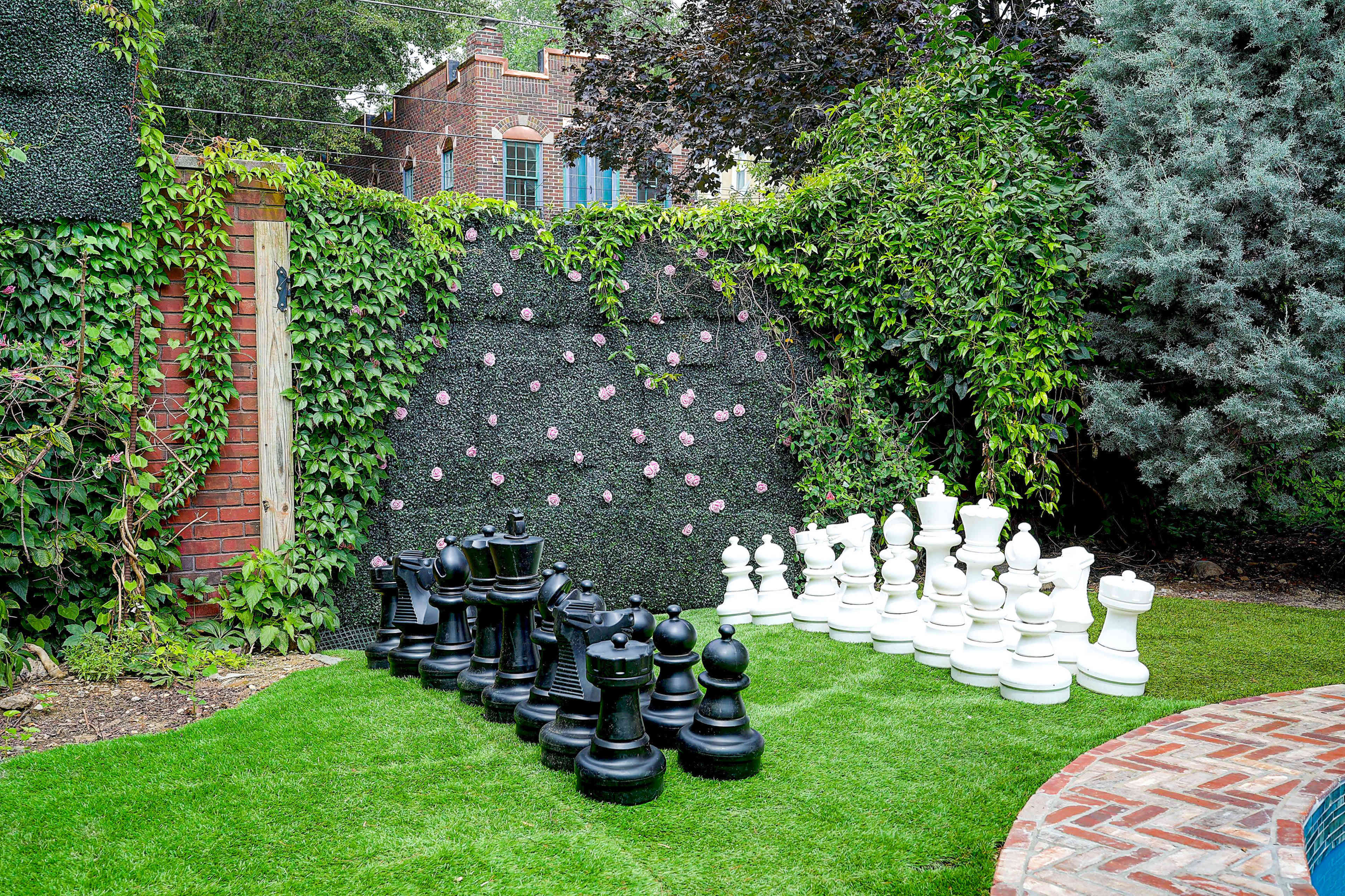 A large outdoor chess set made of black and white pieces is arranged on a grassy area, surrounded by greenery and a brick walkway.