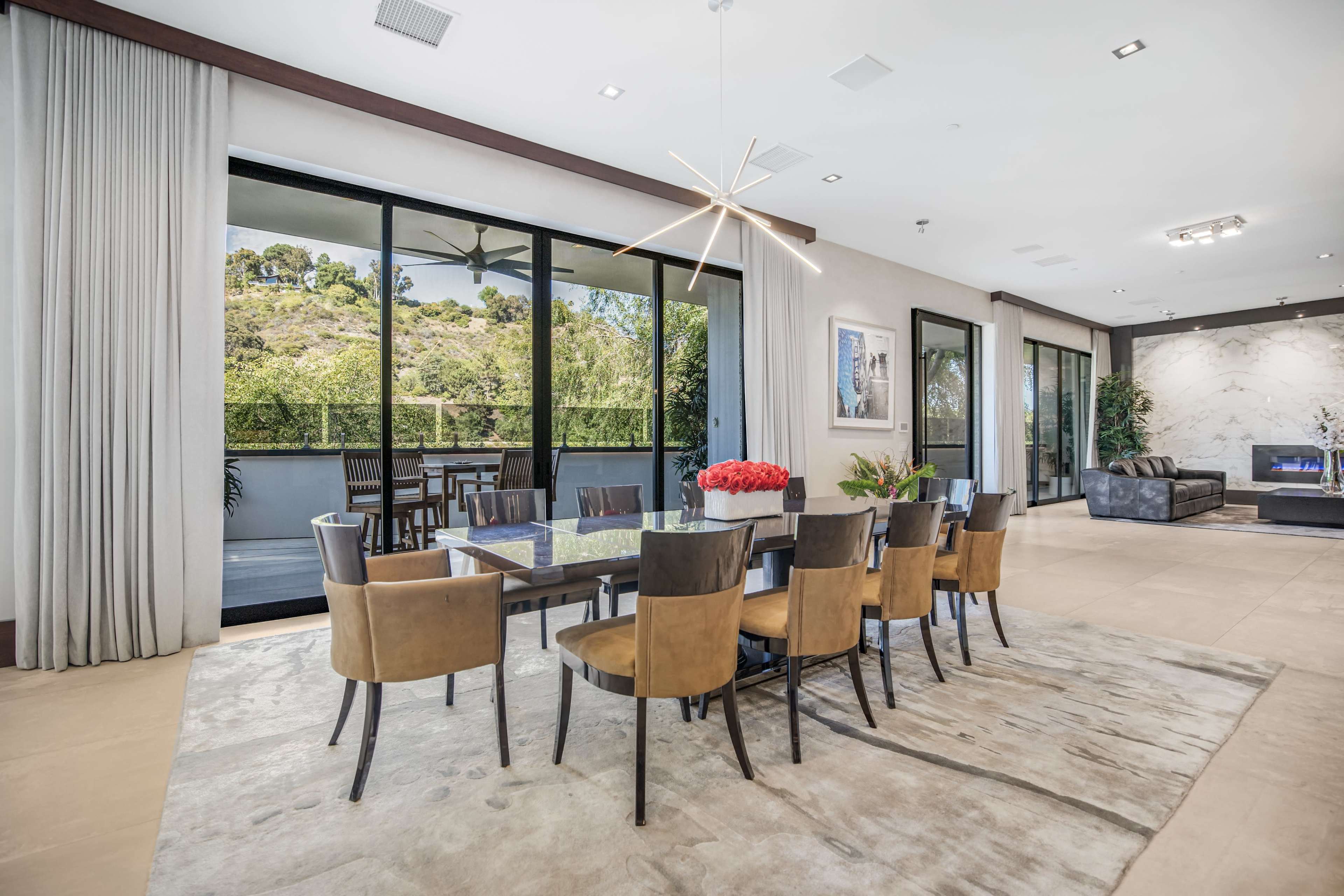 The image shows a modern dining area with a large rectangular table surrounded by eight chairs, overlooking a scenic view through expansive glass doors.