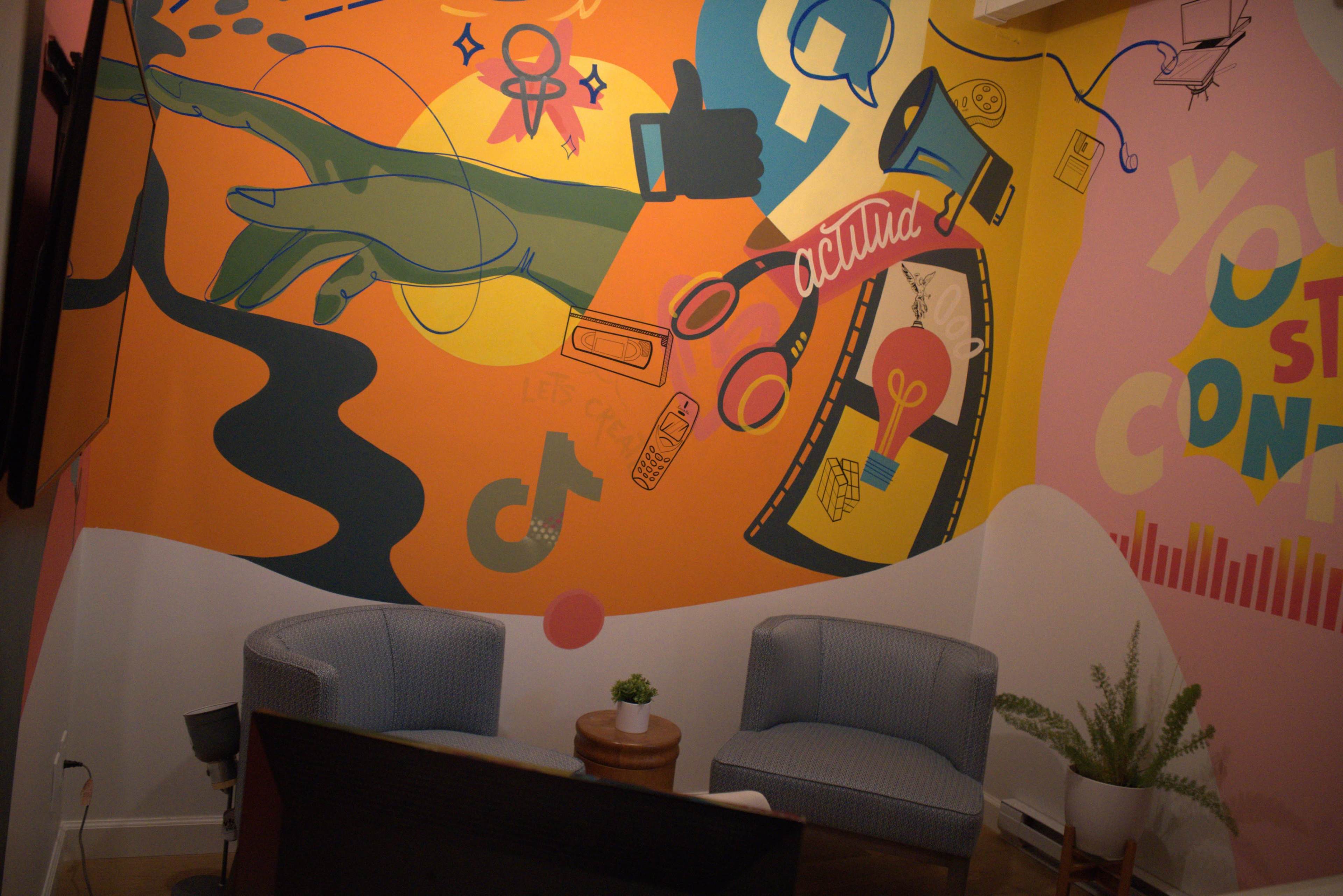 The image shows a vibrant mural featuring colorful designs, including a hand, various symbols, and abstract shapes, against a wall with two chairs and a small plant in front.
