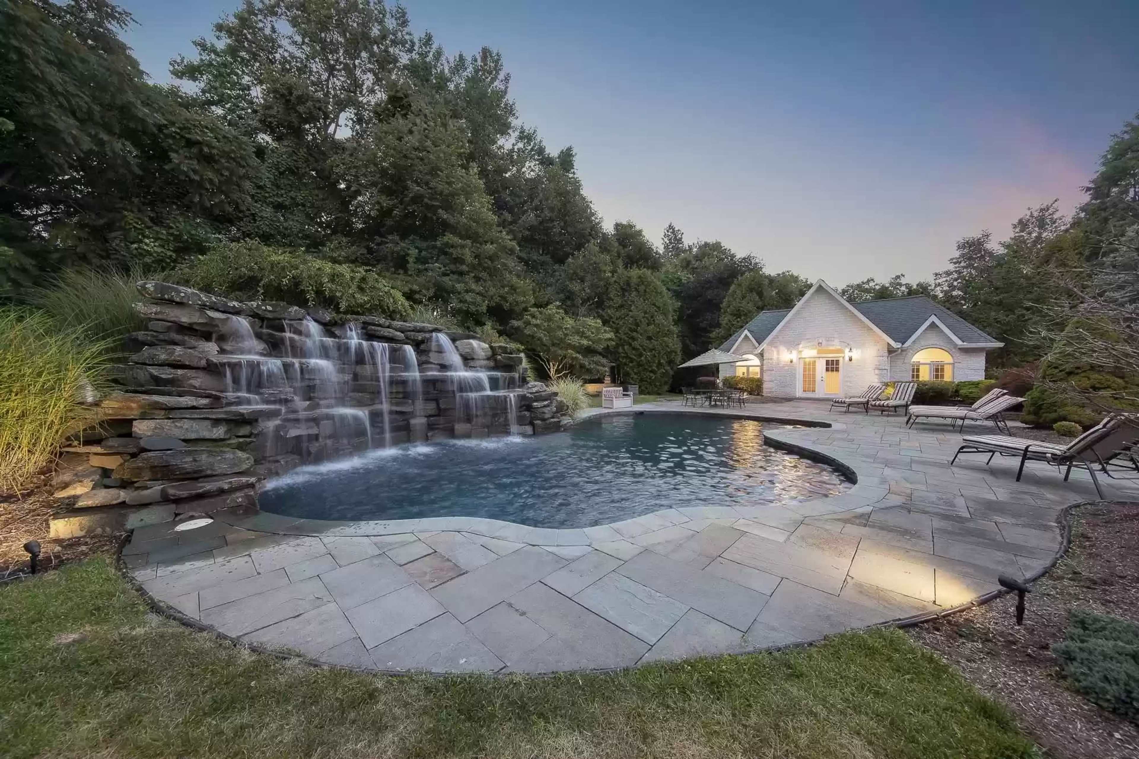 A landscaped backyard featuring a swimming pool with a waterfall surrounded by stone tiles and a house in the background.