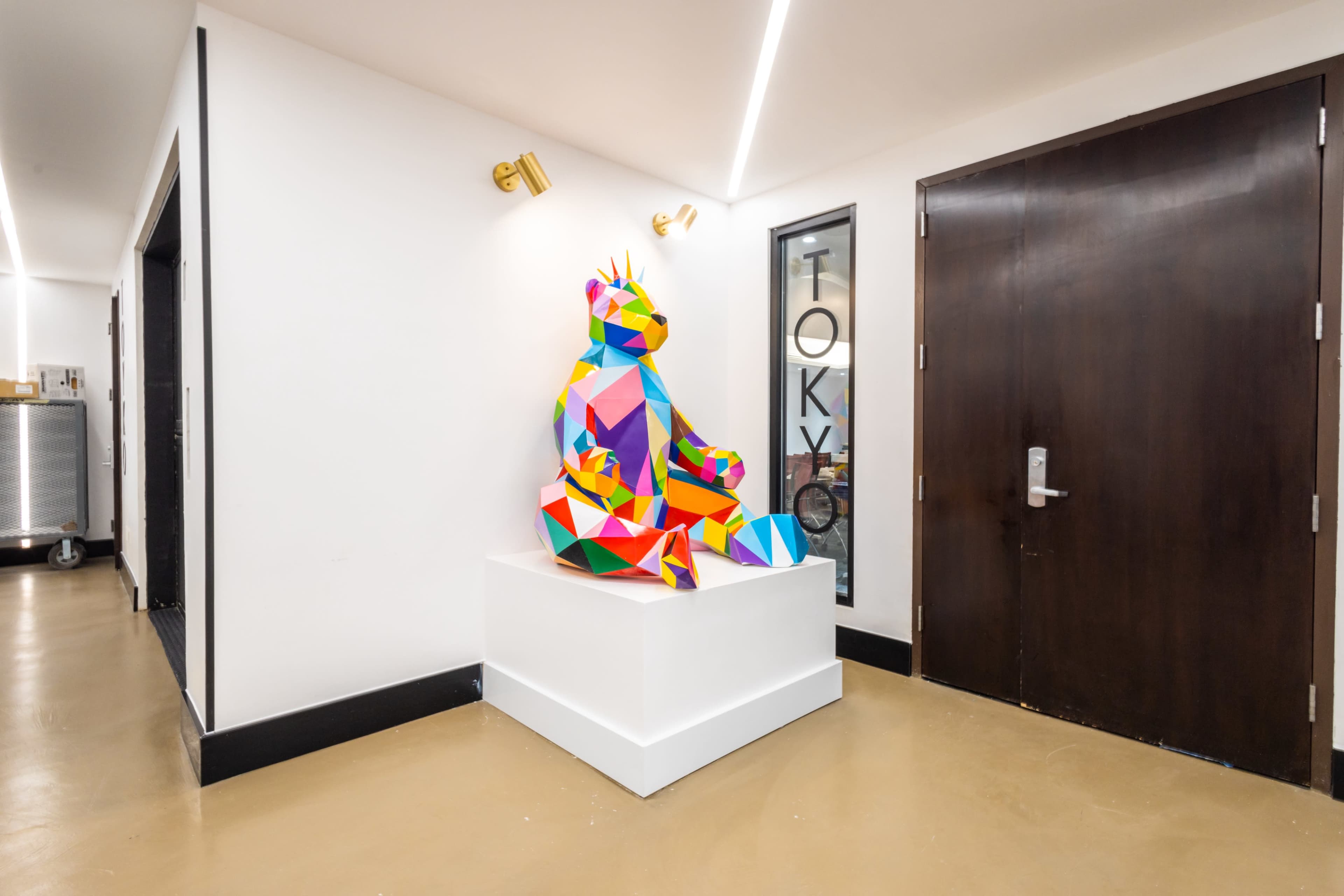 A colorful, geometric bear sculpture sits atop a white pedestal in a hallway near a dark wooden door and glass signage.