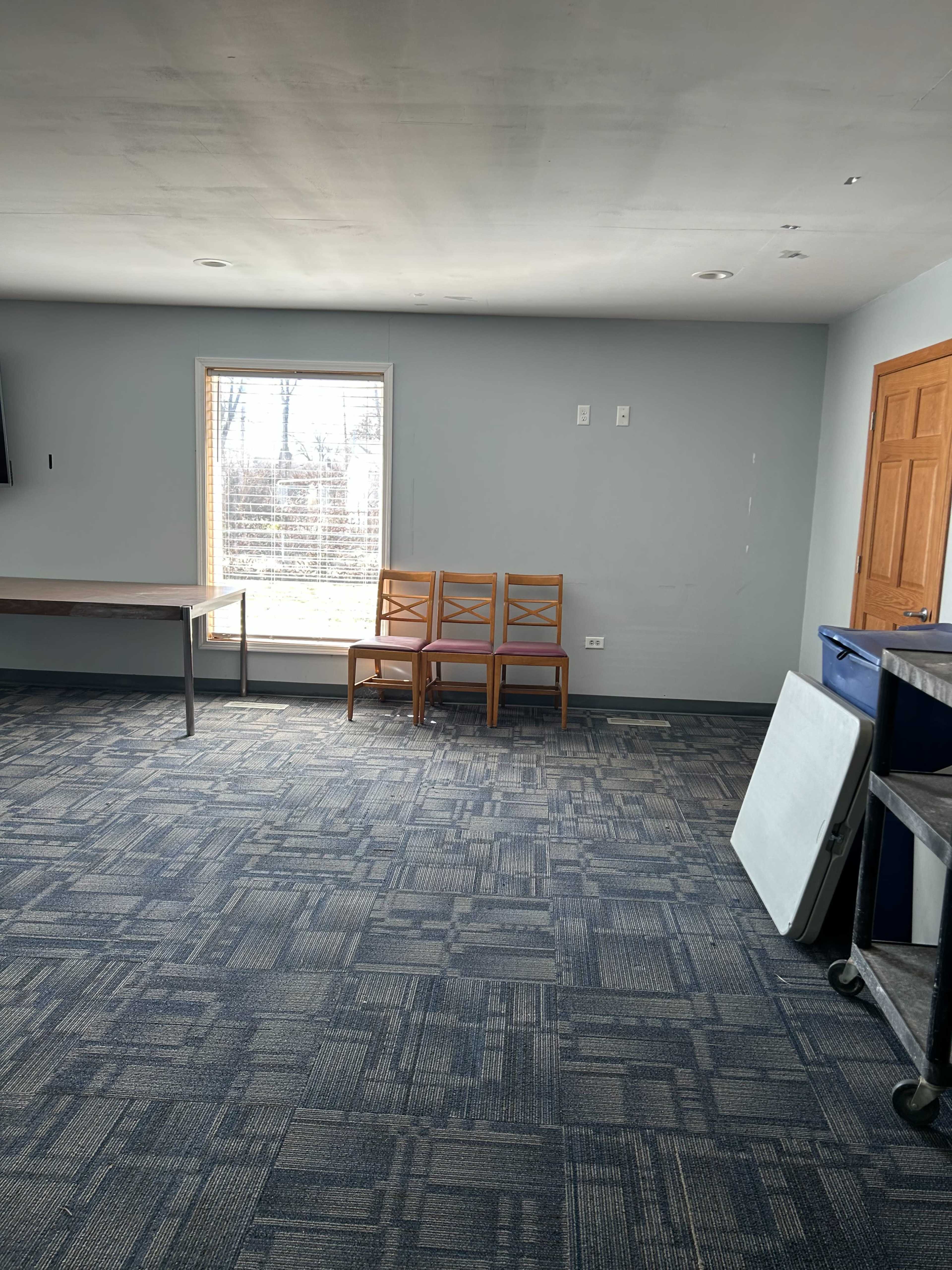 A small, empty room features a window, three wooden chairs against the wall, and a couple of tables positioned around the space.