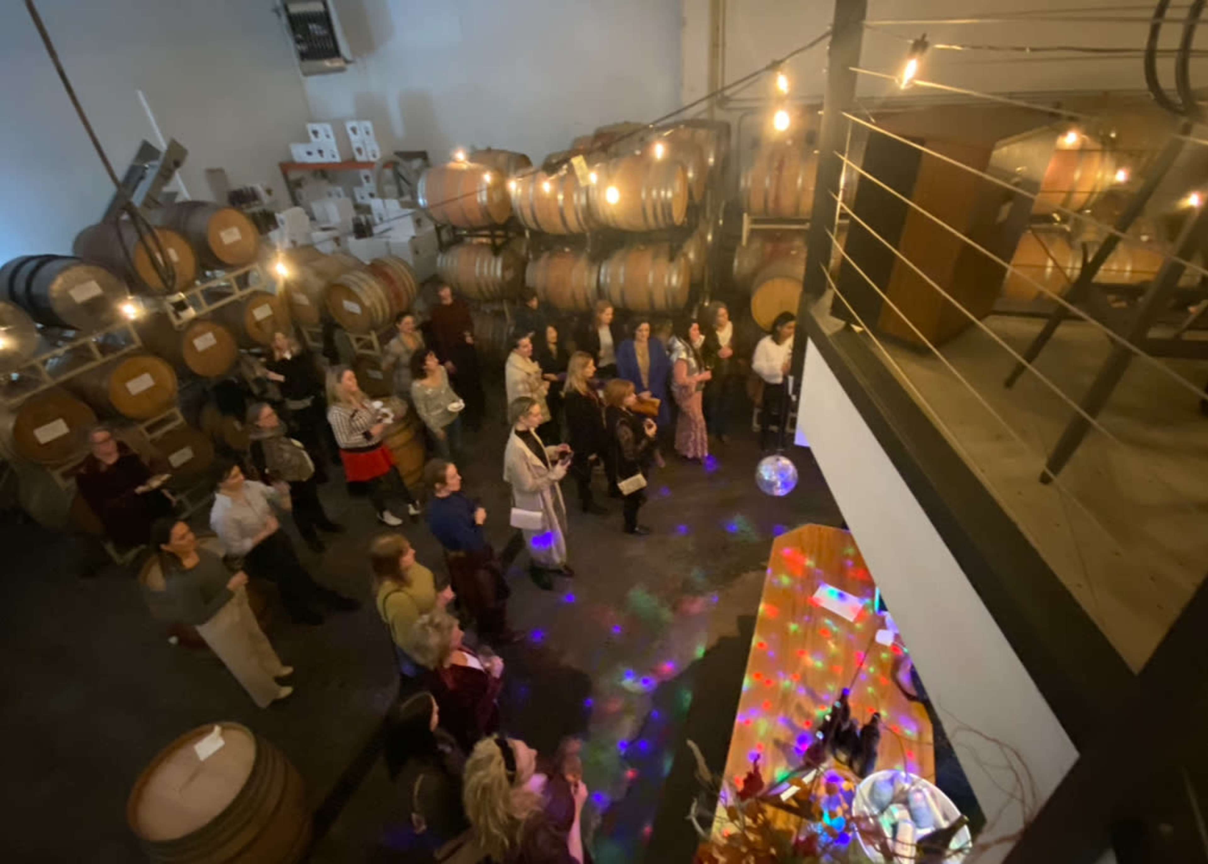 A group of people gathers in a dimly lit winery, surrounded by wooden barrels and colorful lights reflecting off a disco ball.