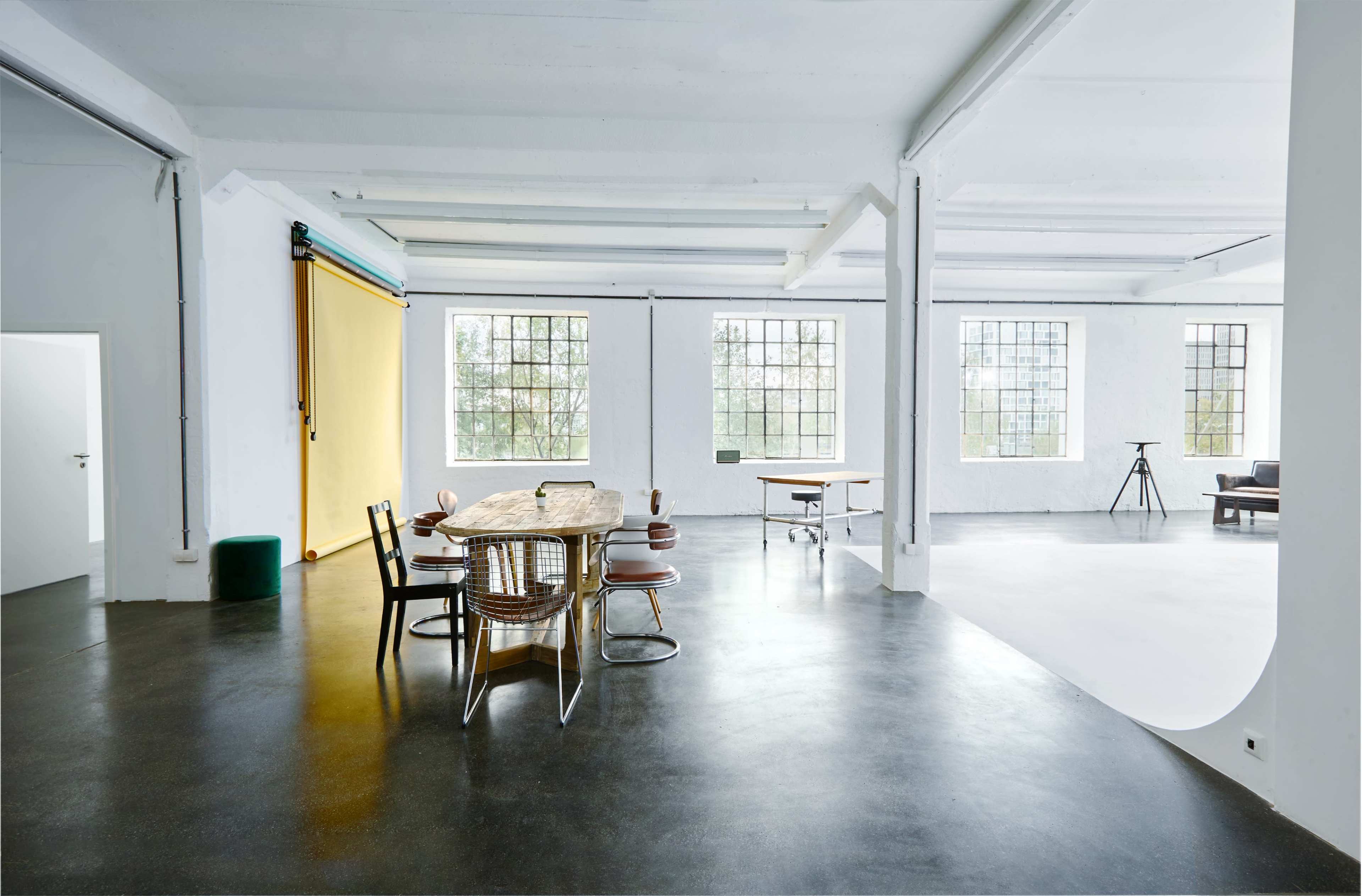 The image shows a spacious, open-plan interior with large windows, featuring a wooden dining table surrounded by chairs, along with a desk and a sitting area.