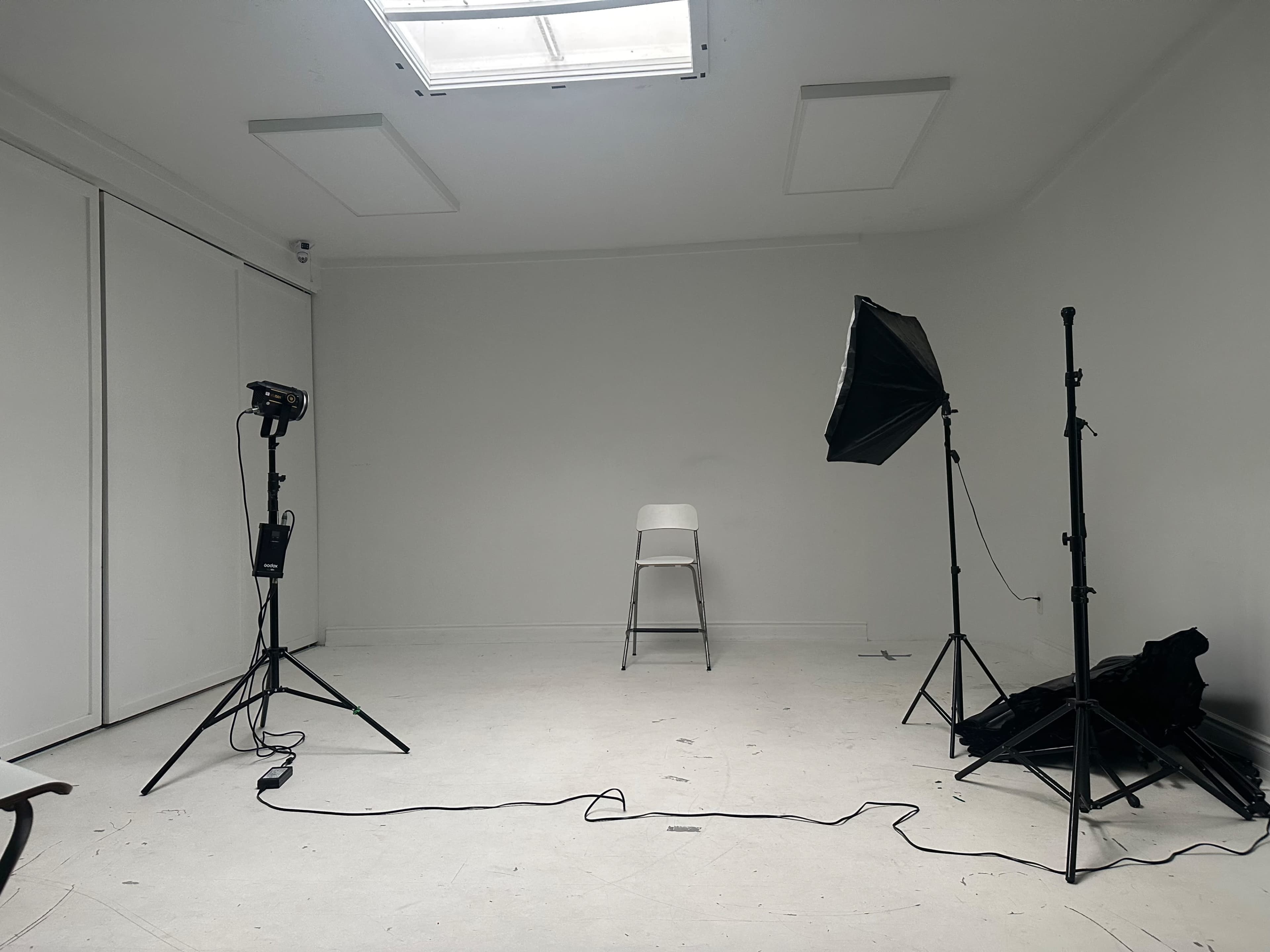 The image shows a minimalistic studio space with a single white chair surrounded by photography equipment and lighting setups.