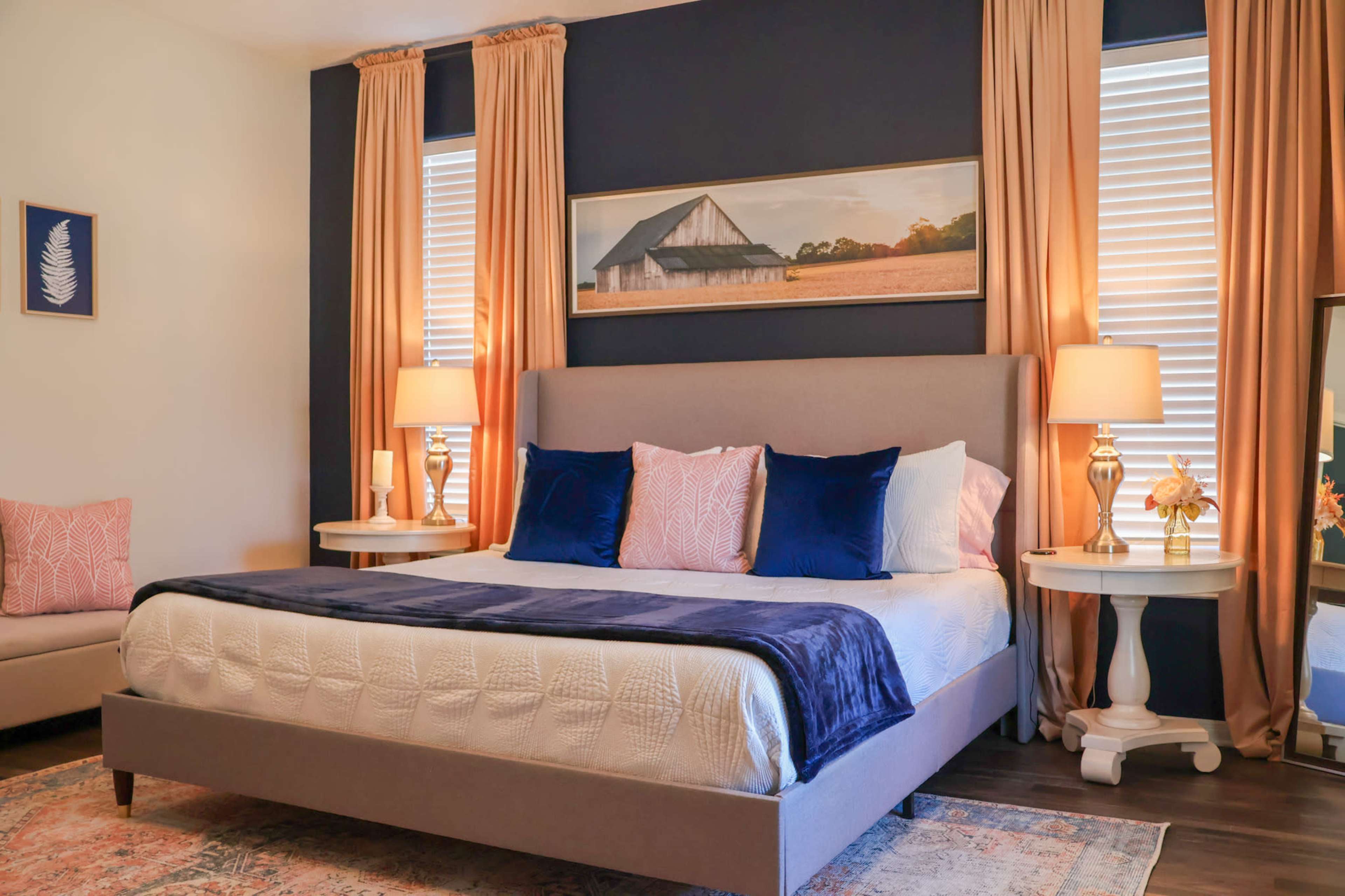 A cozy bedroom featuring a bed with a blue and white color scheme, flanked by two bedside tables with lamps and large windows dressed in long curtains.