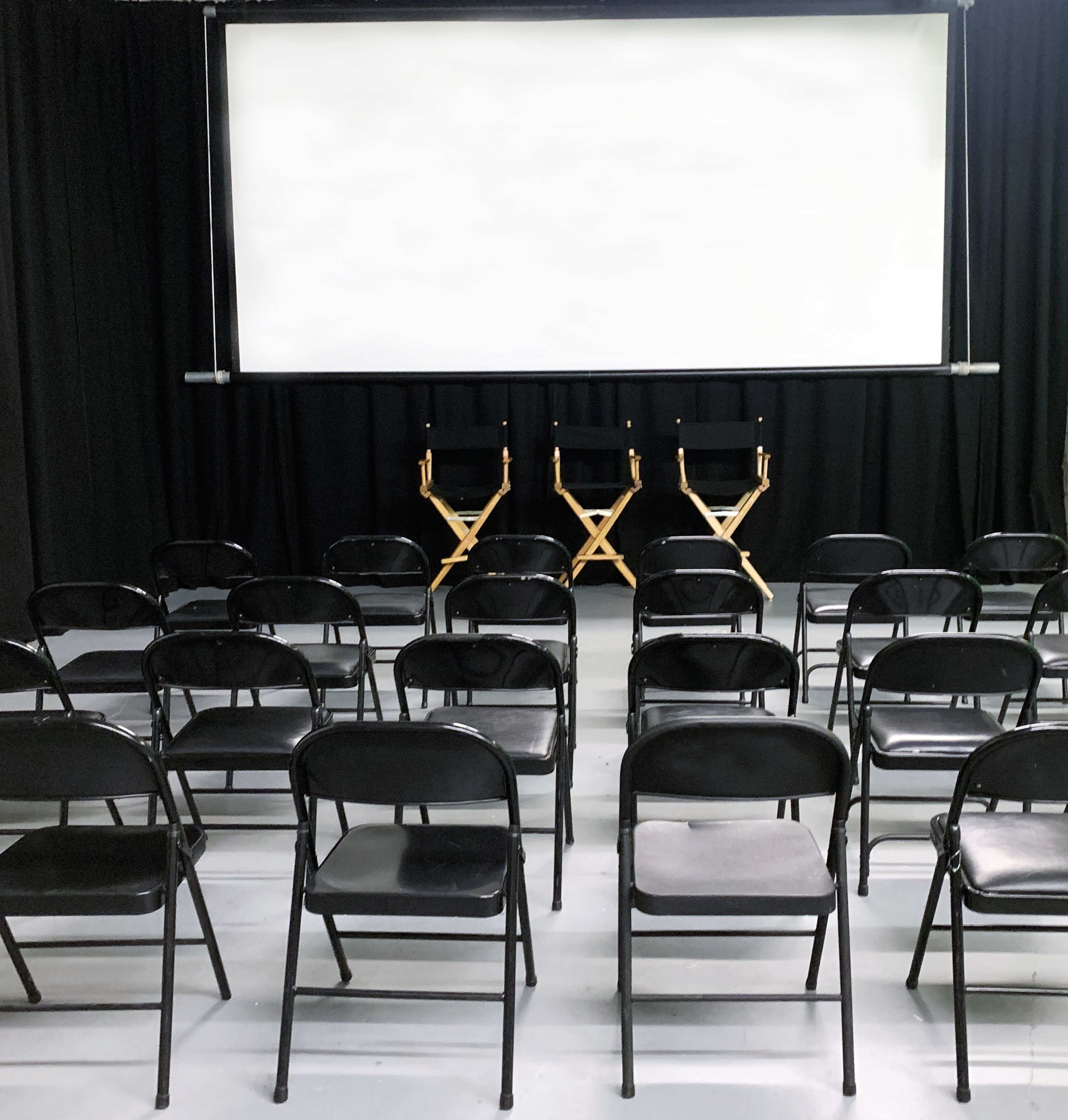 A screening room with black folding chairs arranged in rows facing a blank screen and two director's chairs on stage.
