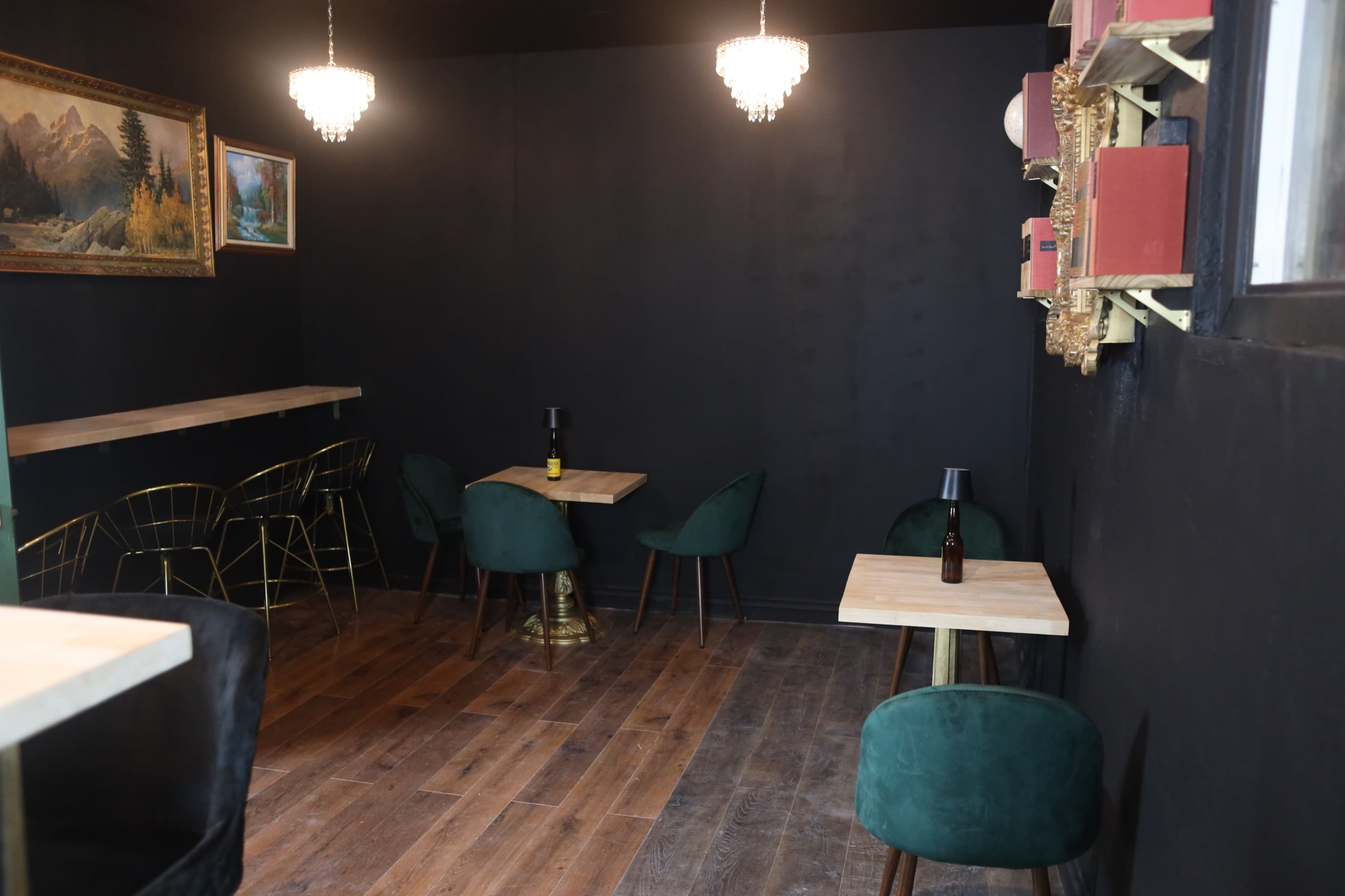 The image features a dimly lit café space with dark walls, wooden flooring, and a few small tables surrounded by green chairs, along with decorative chandeliers hanging from the ceiling.