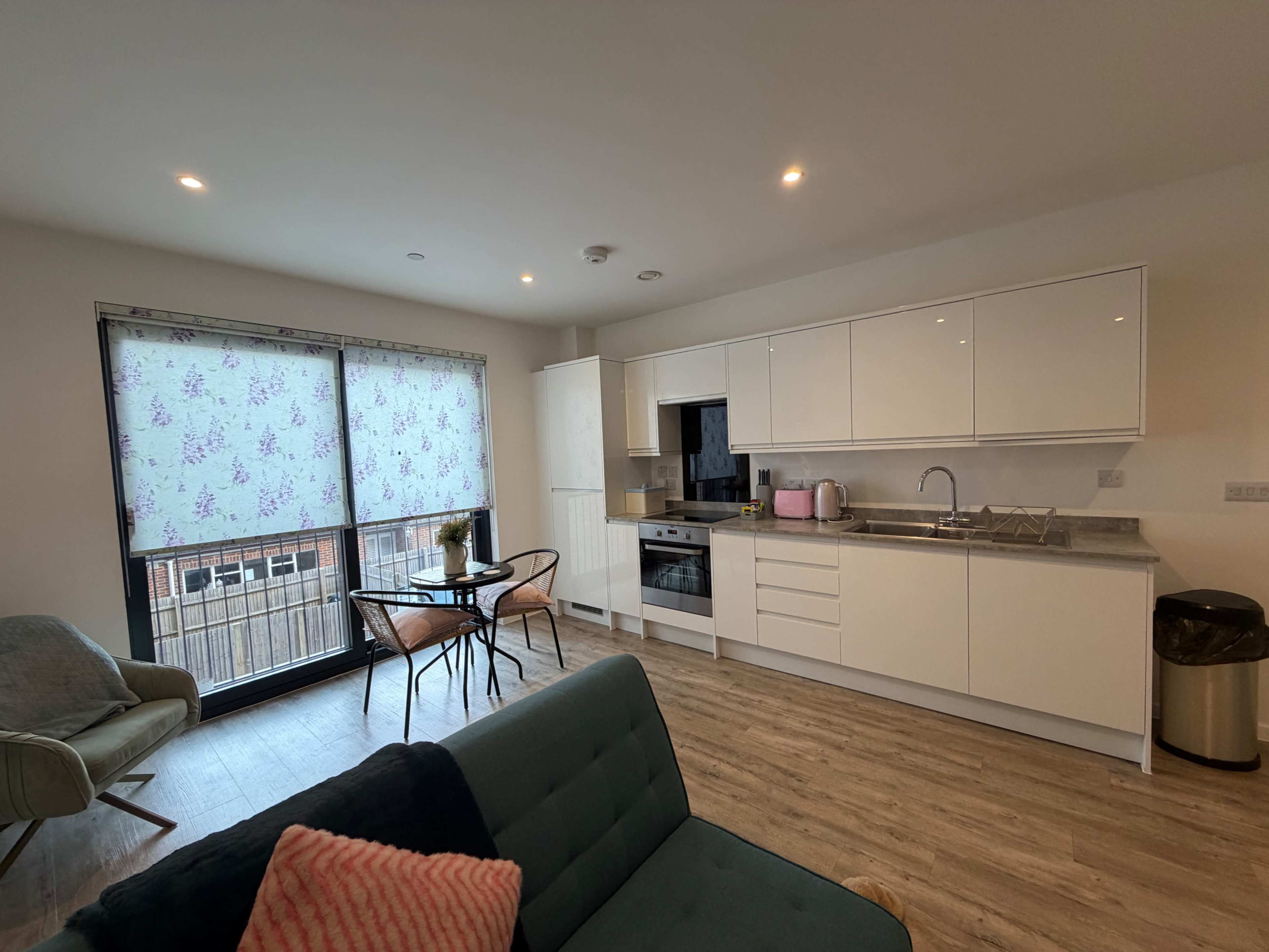 The image shows a modern kitchen and living area with a small dining table and chairs next to a large window covered by floral curtains.