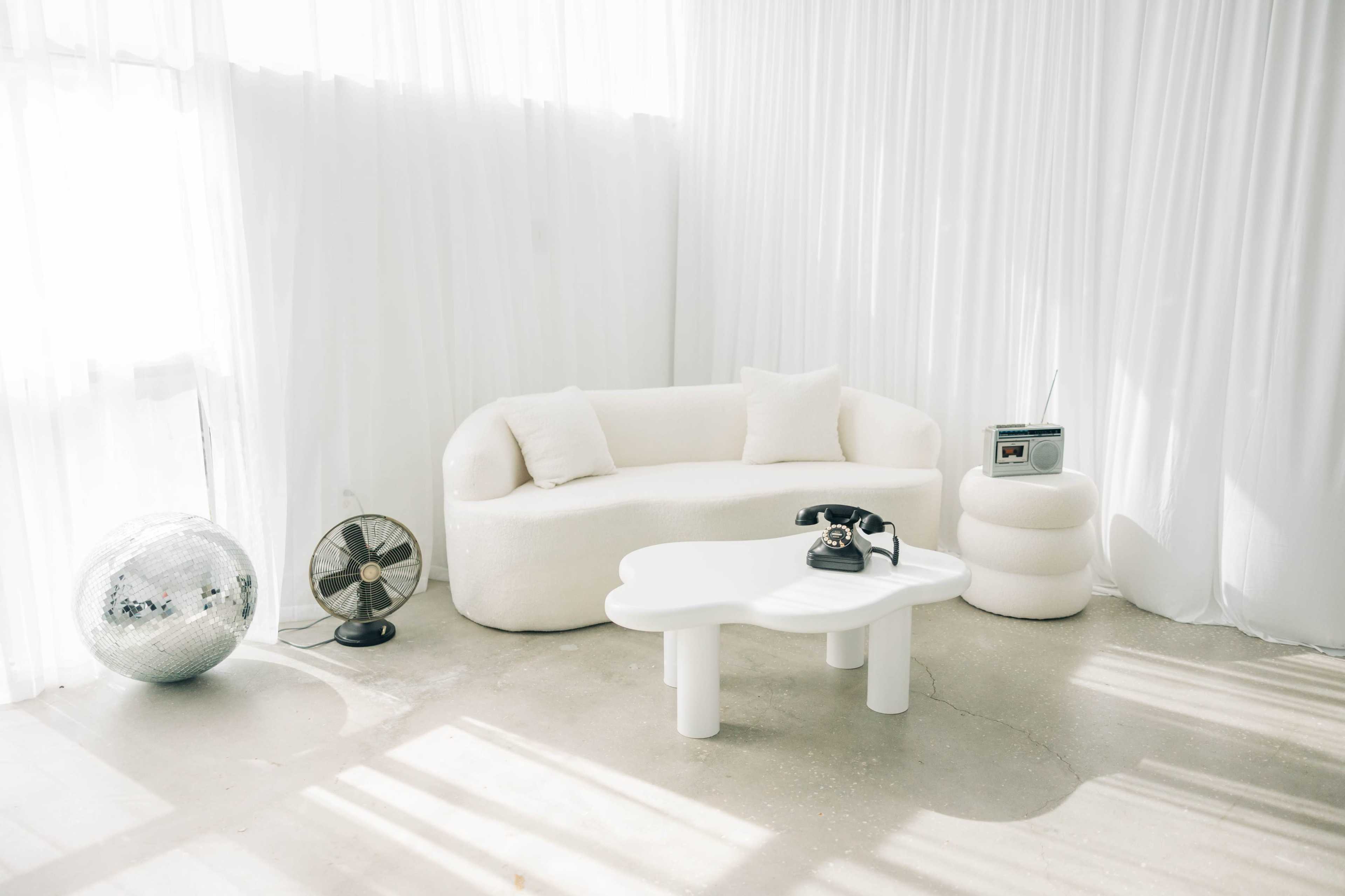 The scene features a softly curved white sofa, a round coffee table, a black rotary phone, a vintage radio, a fan, and a disco ball, all set against sheer white drapery in a bright room.