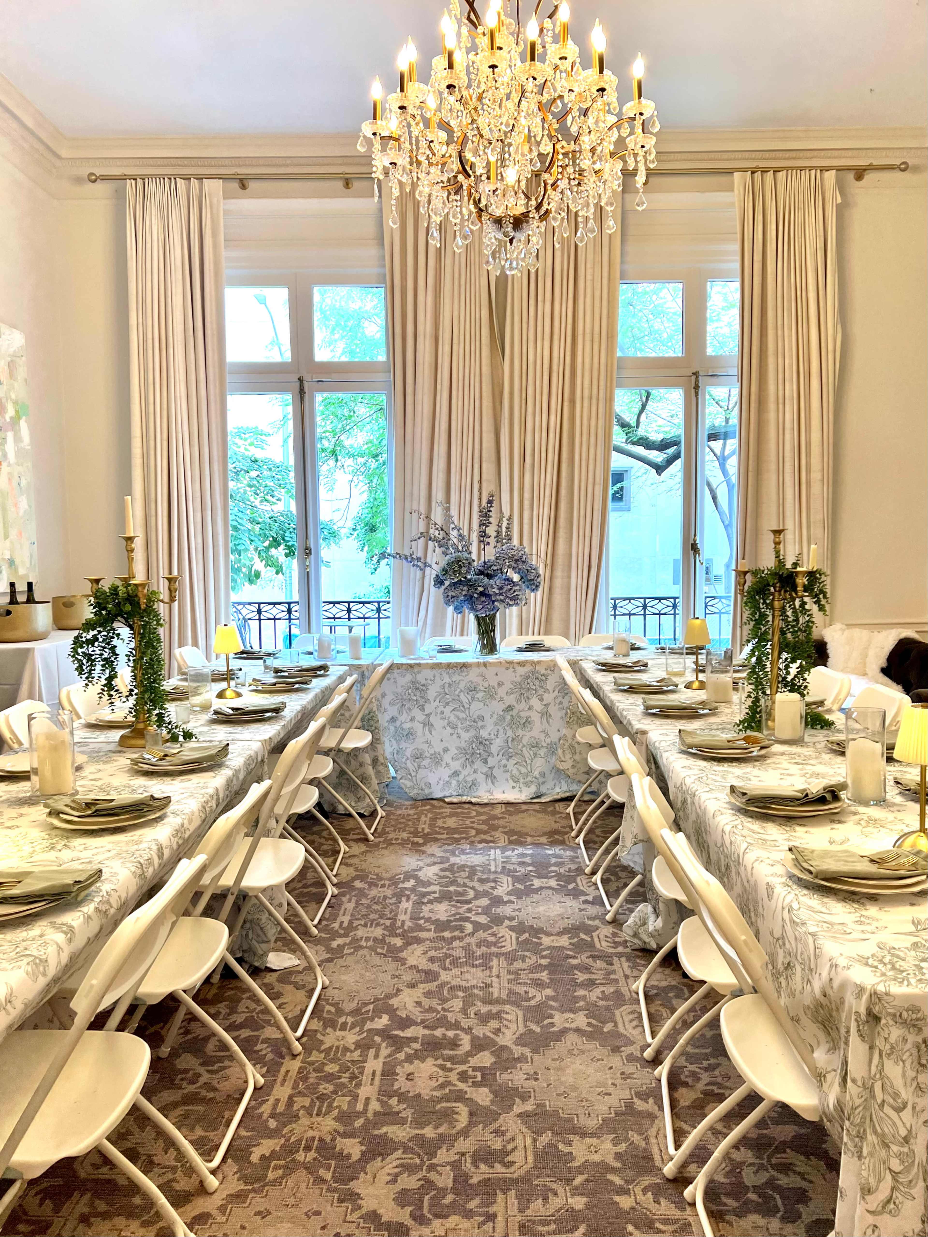 A long dining table is set with decorative tableware and flowers, under a chandelier in a bright, elegant room with large windows.
