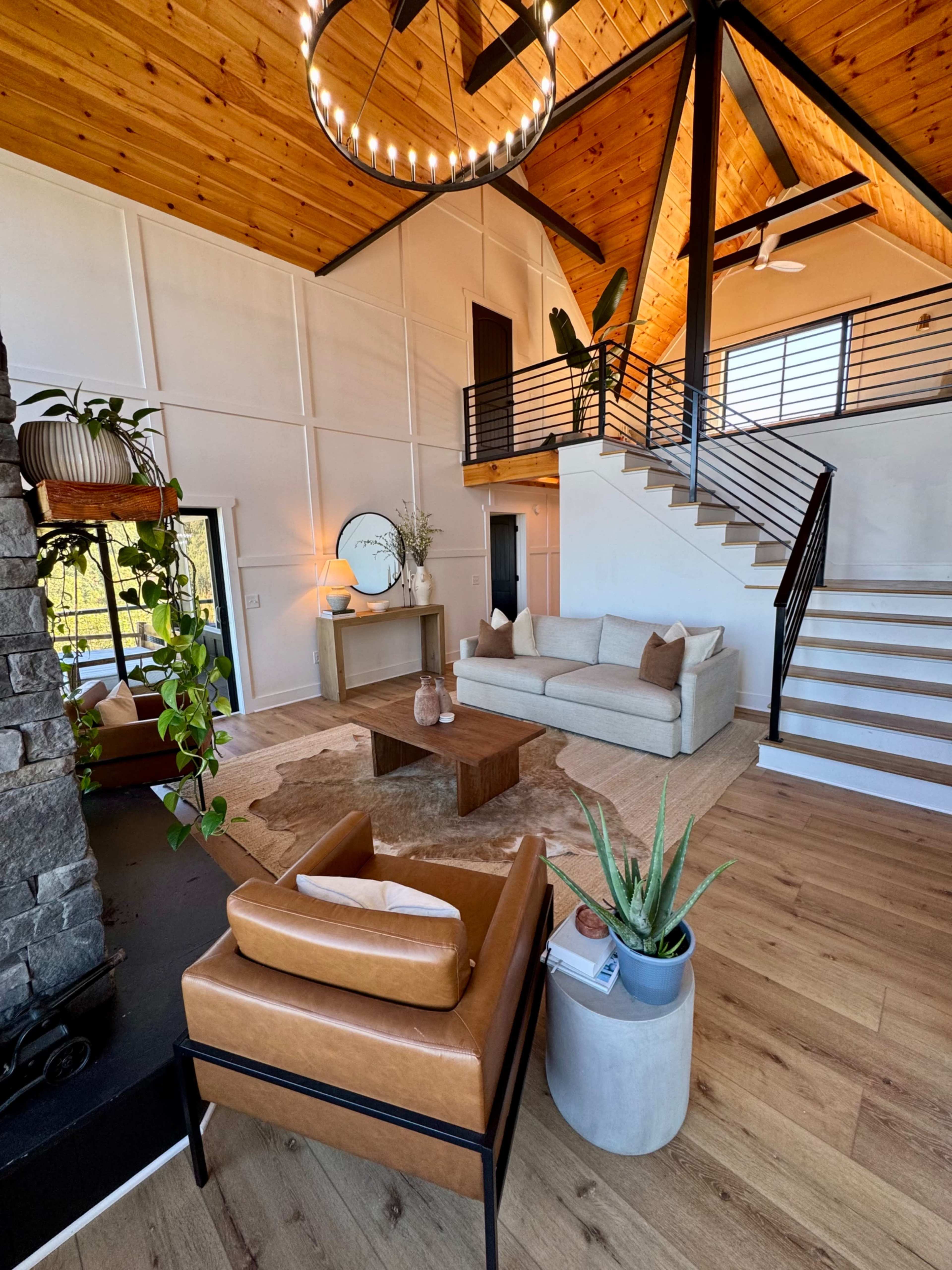 The image shows a modern living room featuring a high ceiling, large windows, a staircase with a black railing, a beige sofa, wooden flooring, and decorative plants.