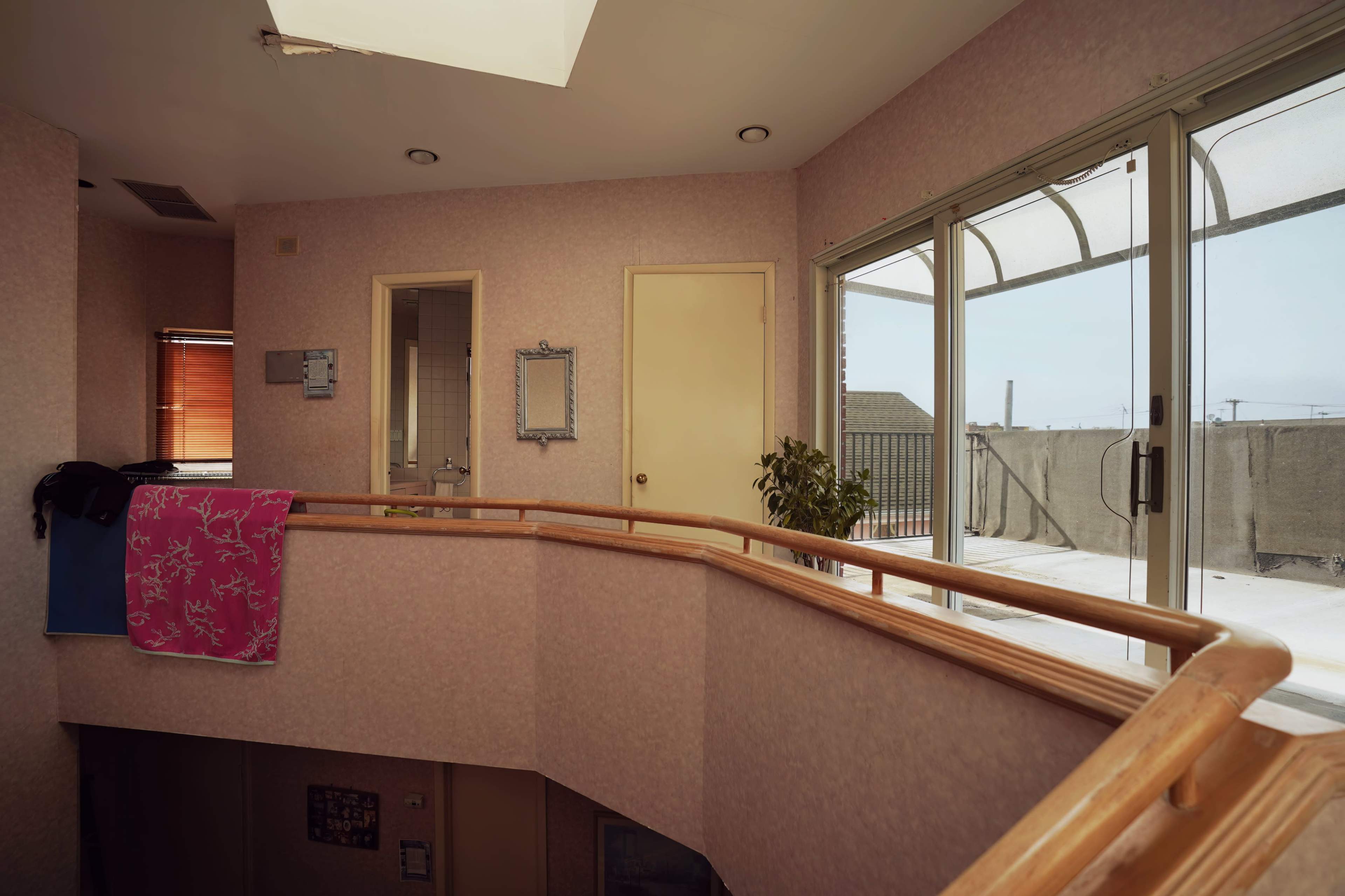 The image shows an interior space with a wooden railing overlooking a patio door, leading to an outdoor area with a small plant in the corner and a pink towel hanging on the railing.
