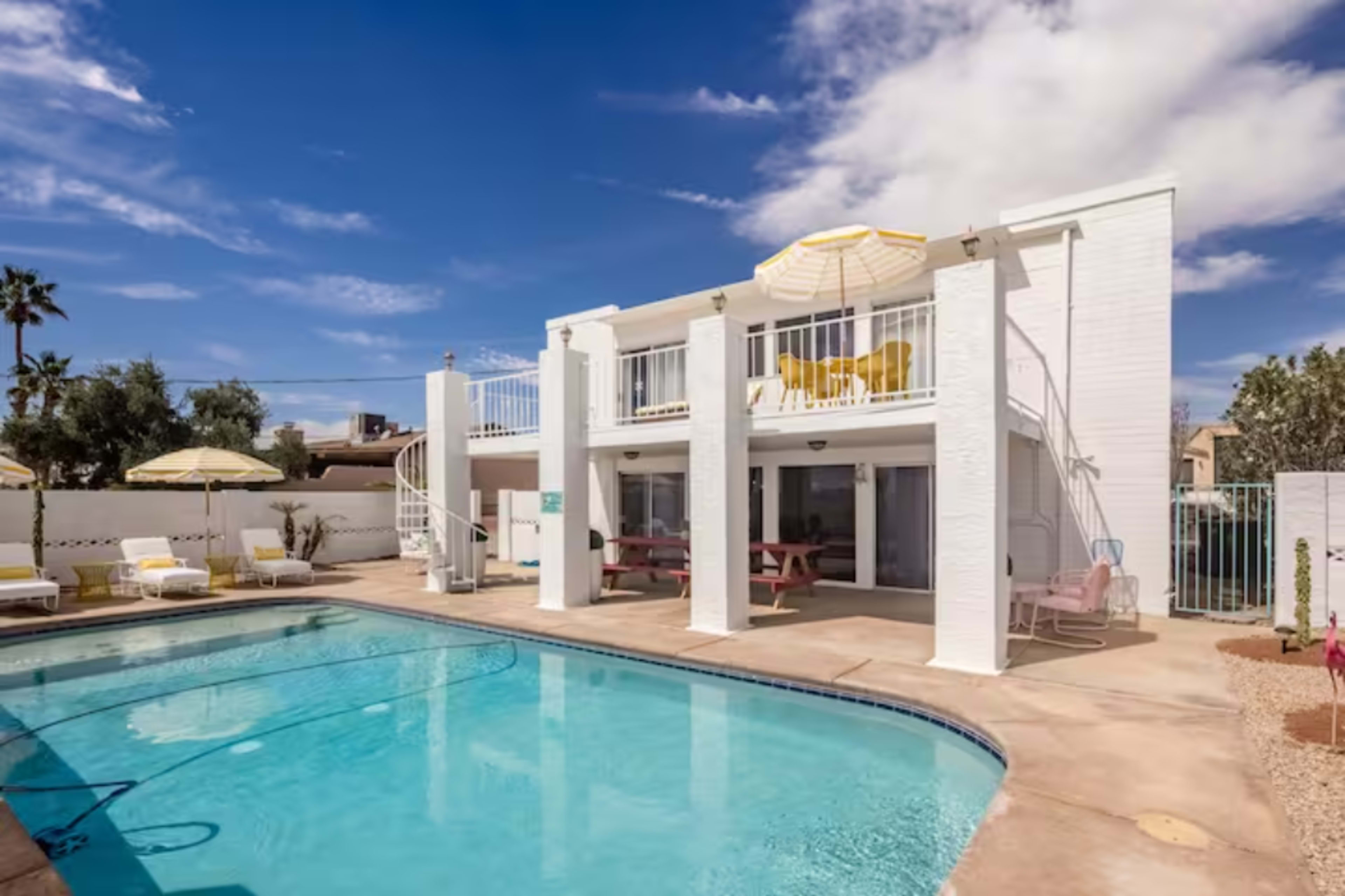 A white two-story house overlooks a swimming pool with lounge chairs and umbrellas nearby.