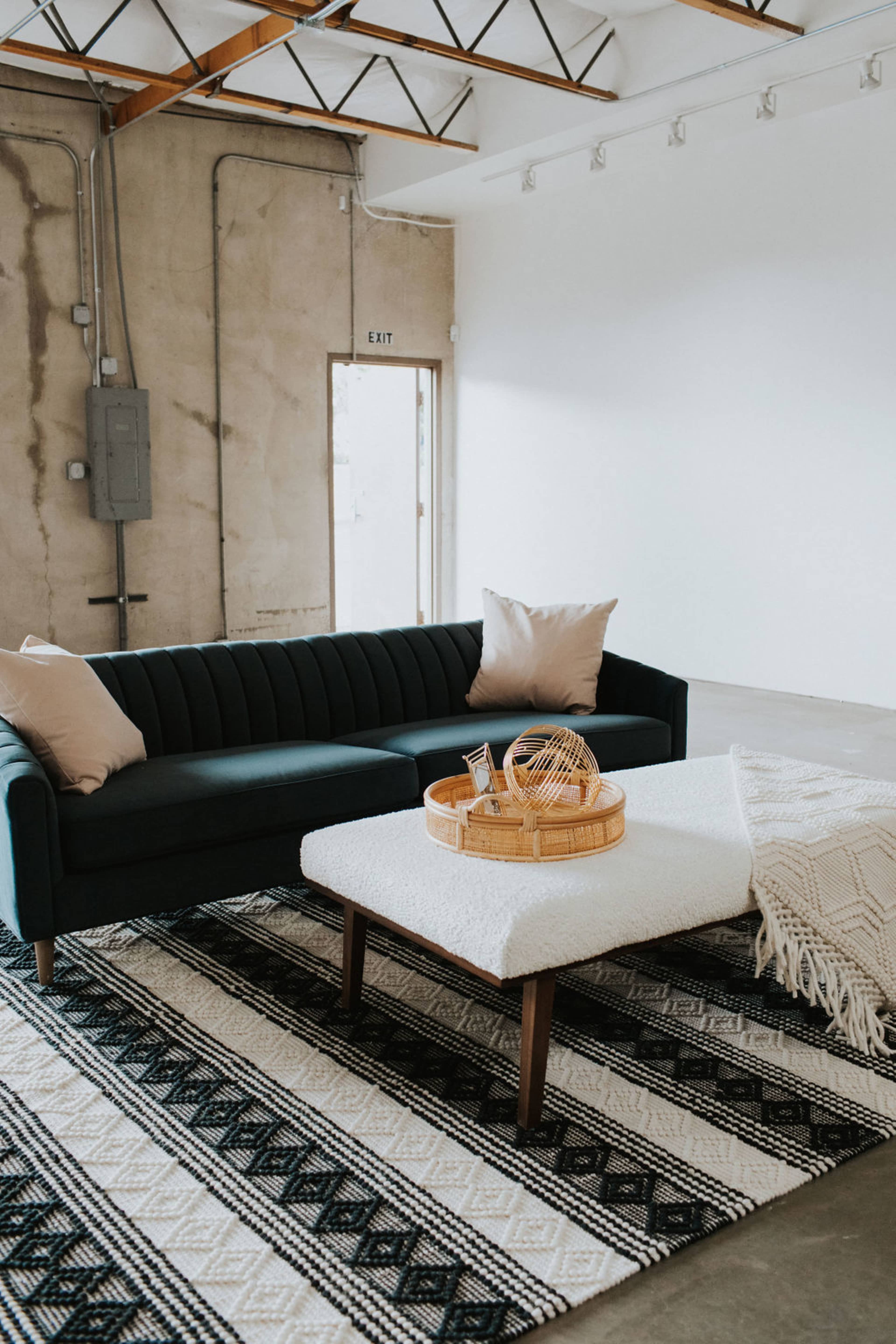 A modern living room featuring a dark green upholstered sofa with pillows, a wooden coffee table with a woven tray, and a patterned rug on a concrete floor.