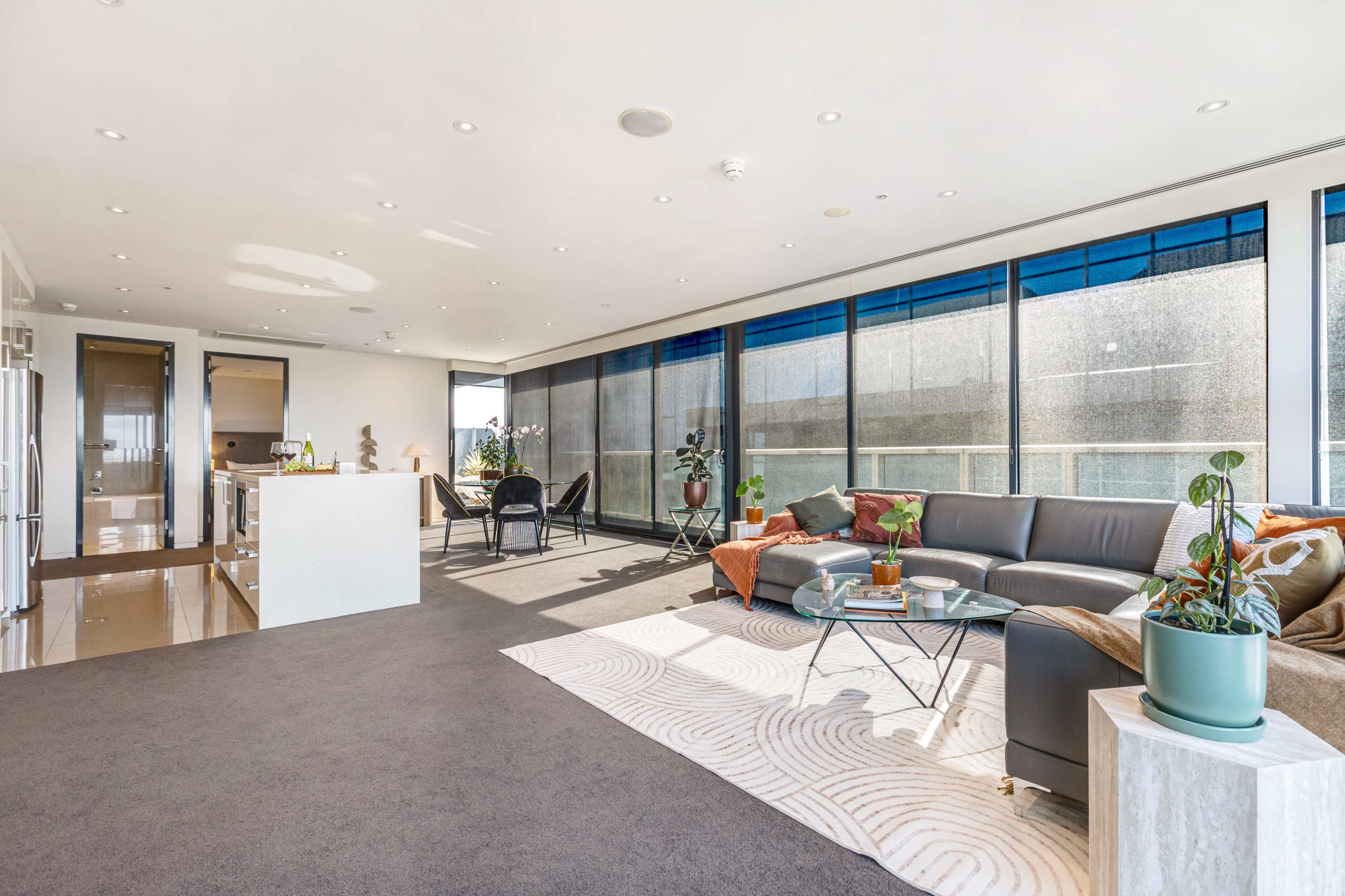 The image shows a modern, open-concept living space with large windows, a comfortable couch, and a small dining area.
