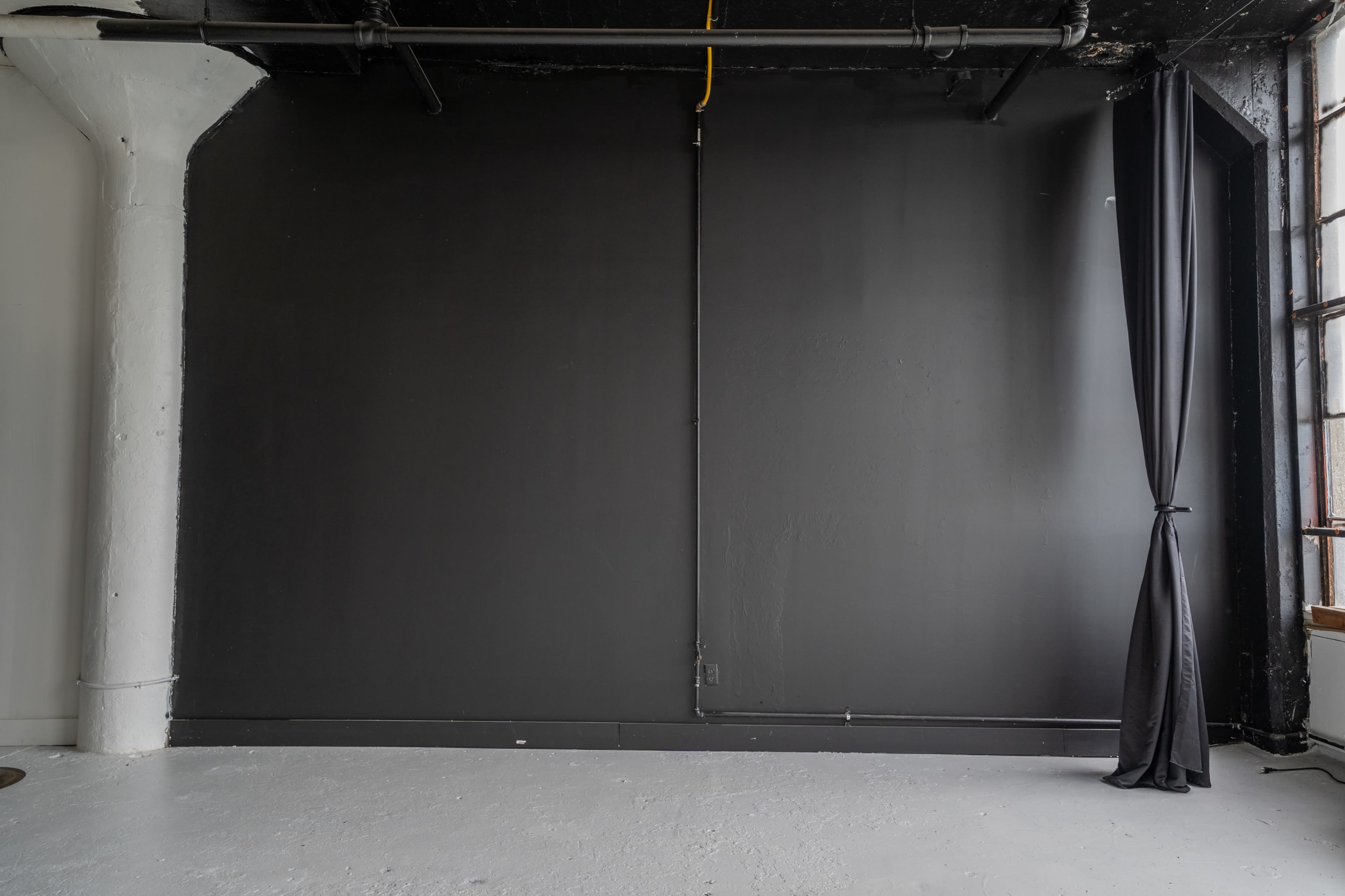 A spacious interior with a black wall, partially covered by a black curtain, and exposed pipes running along the ceiling.