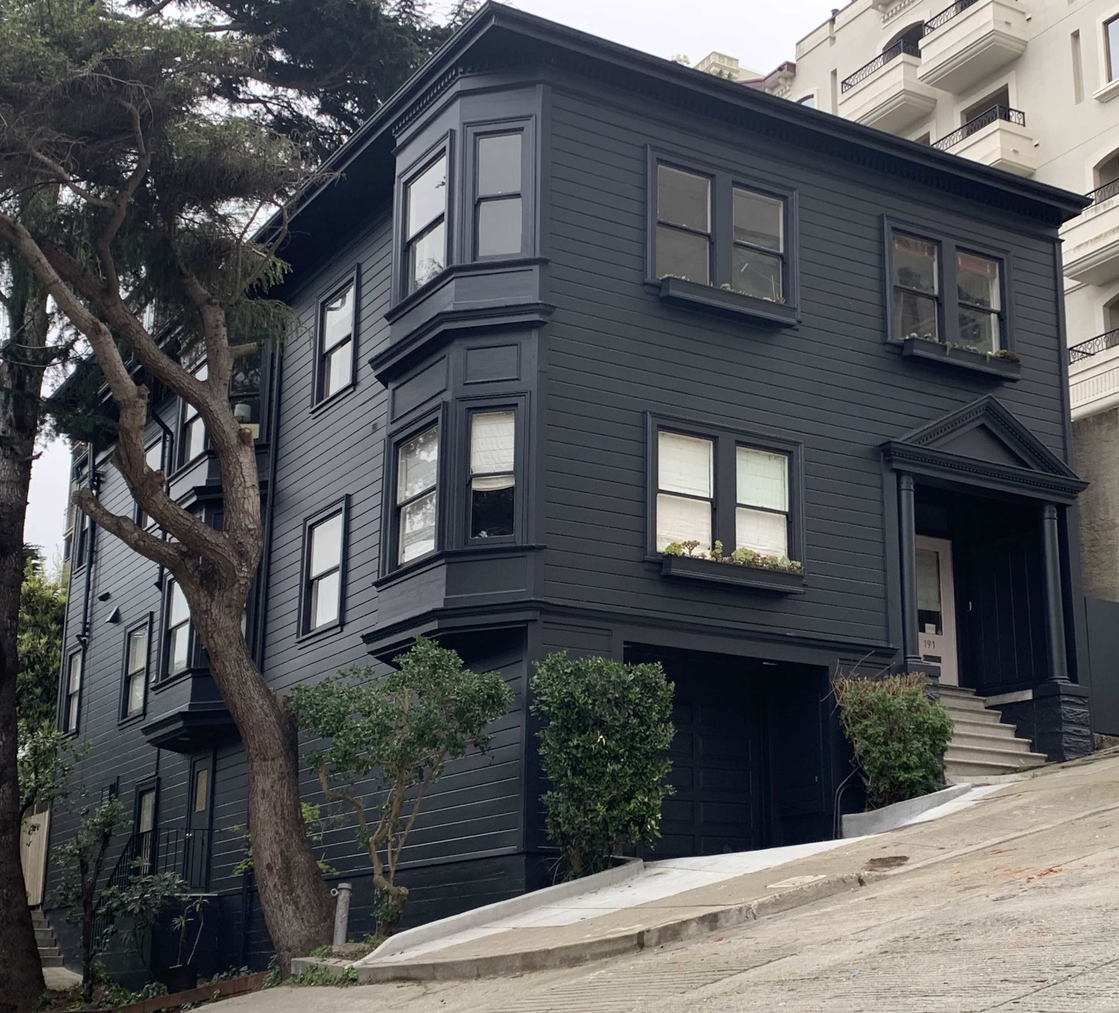 A three-story black wooden house with bay windows sits on a steeply sloped street, surrounded by greenery.