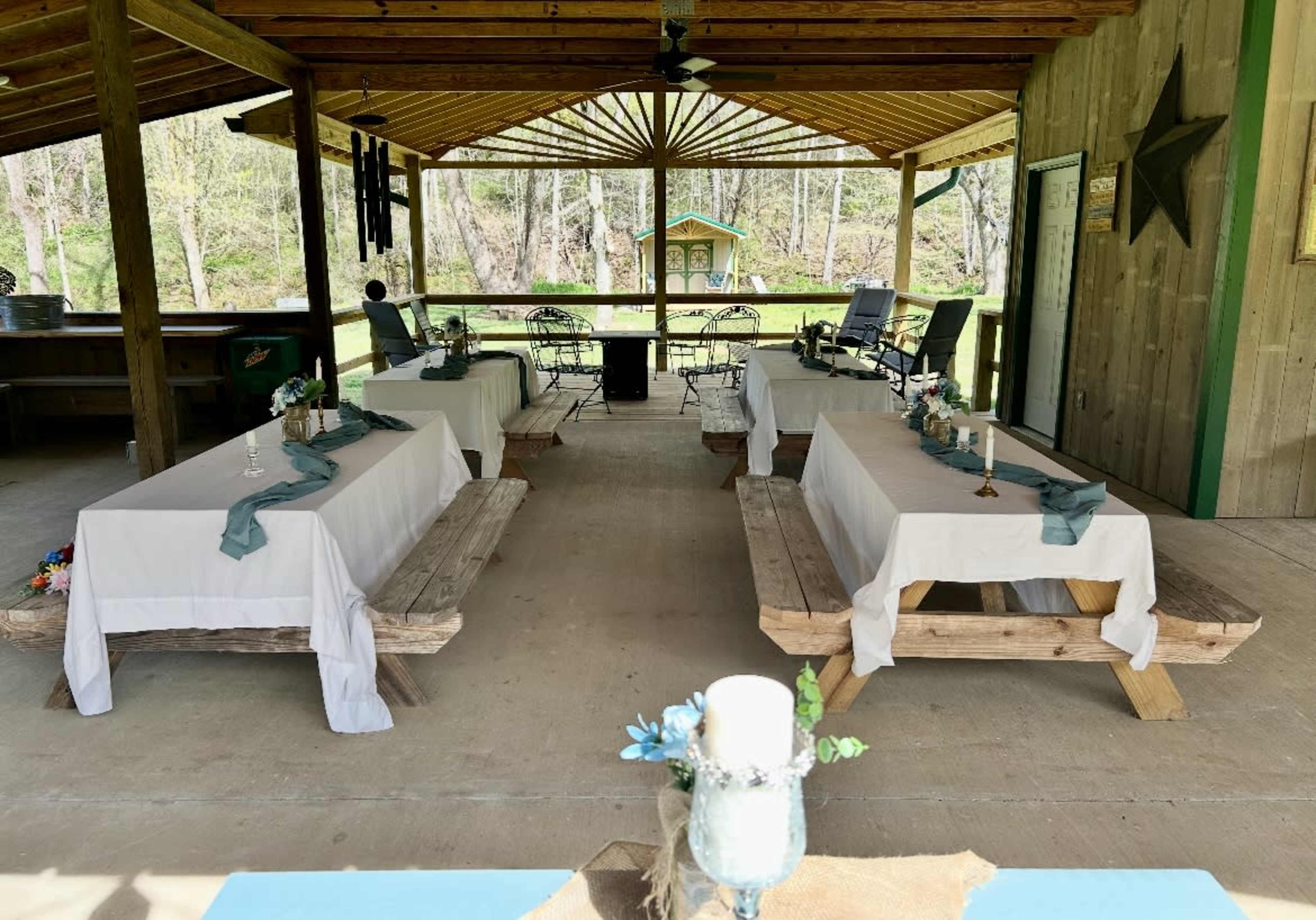 A spacious outdoor venue features several long tables covered with white tablecloths and decorated with teal runners, set up beneath a wooden pavilion.