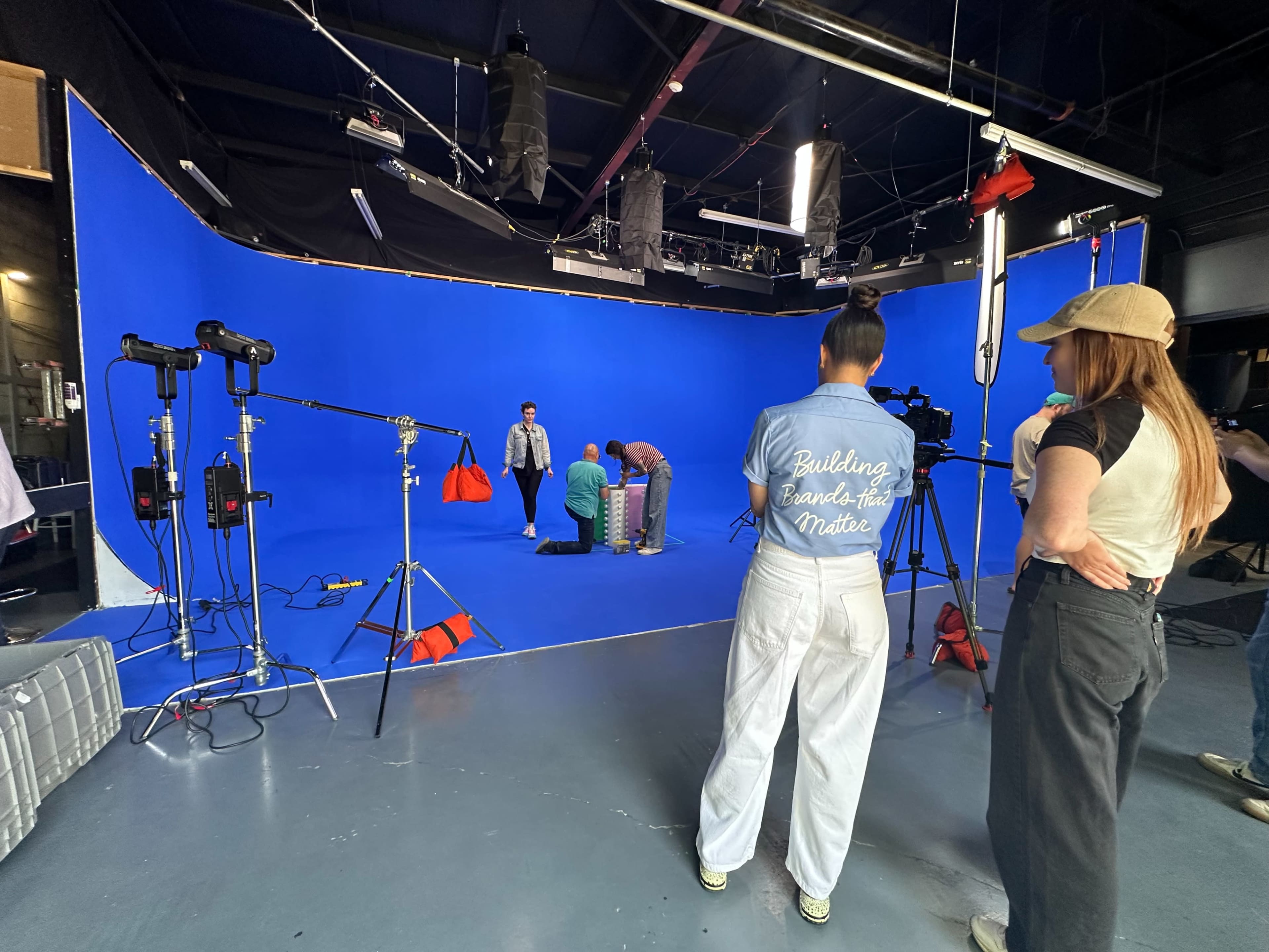 A group of people is preparing a photoshoot in a studio with a blue backdrop, featuring lighting equipment and a model adjusting items on a platform.