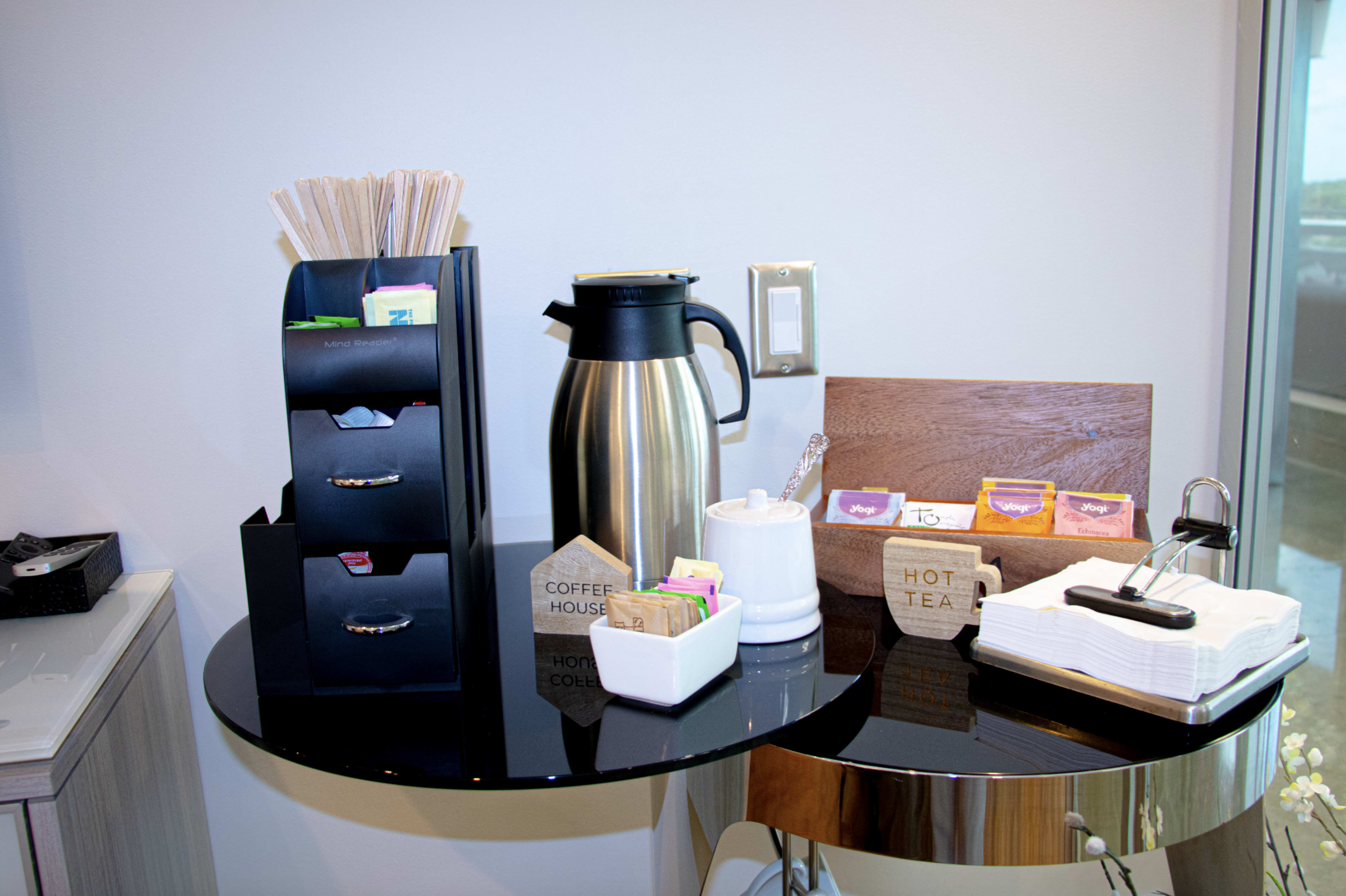 A coffee station is displayed on a black surface, featuring a thermos, various tea bags, sugar, and stirrers.