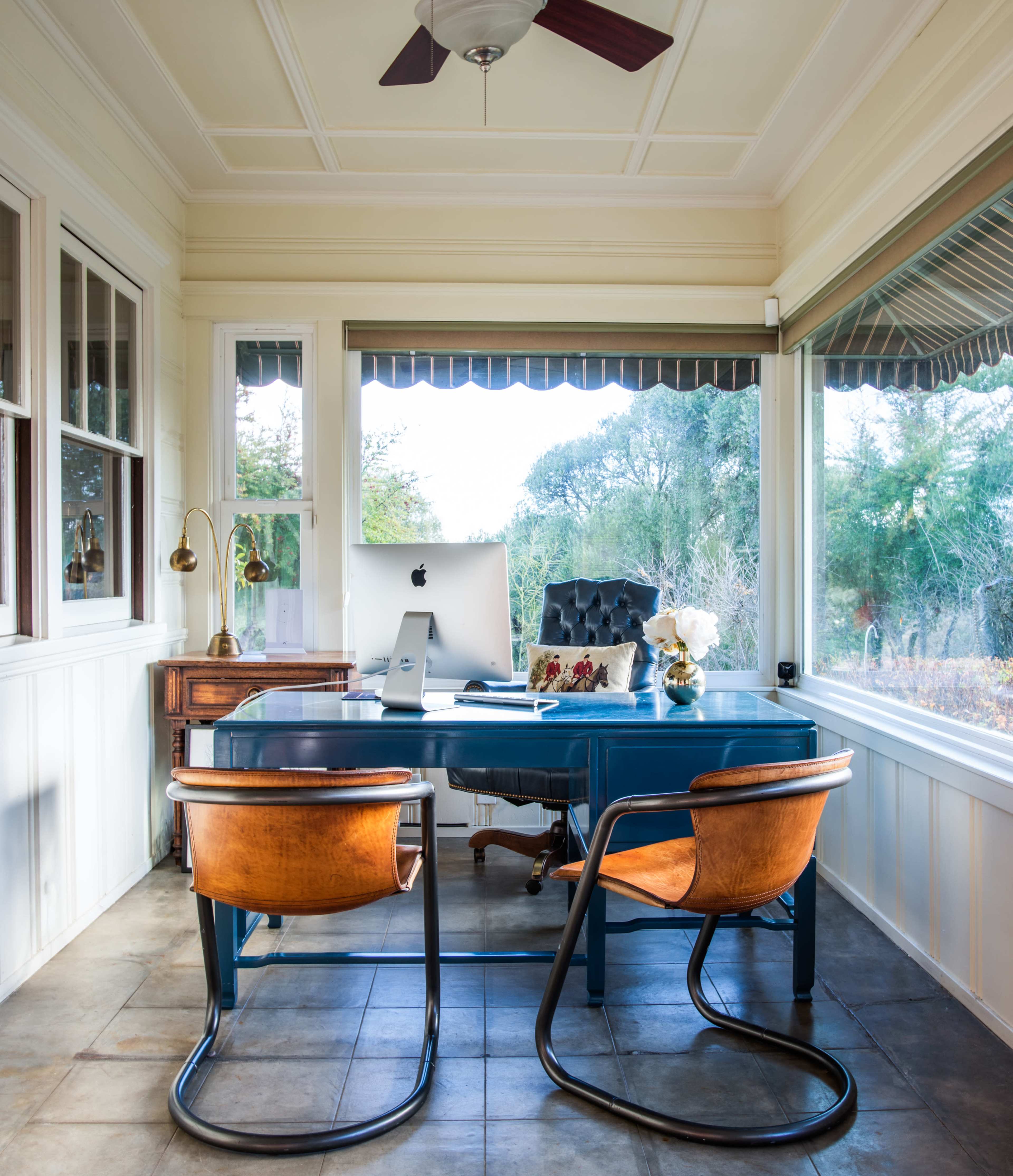 A stylish home office features a blue desk with an iMac, two curved wooden chairs, and large windows showcasing a view of greenery outside.