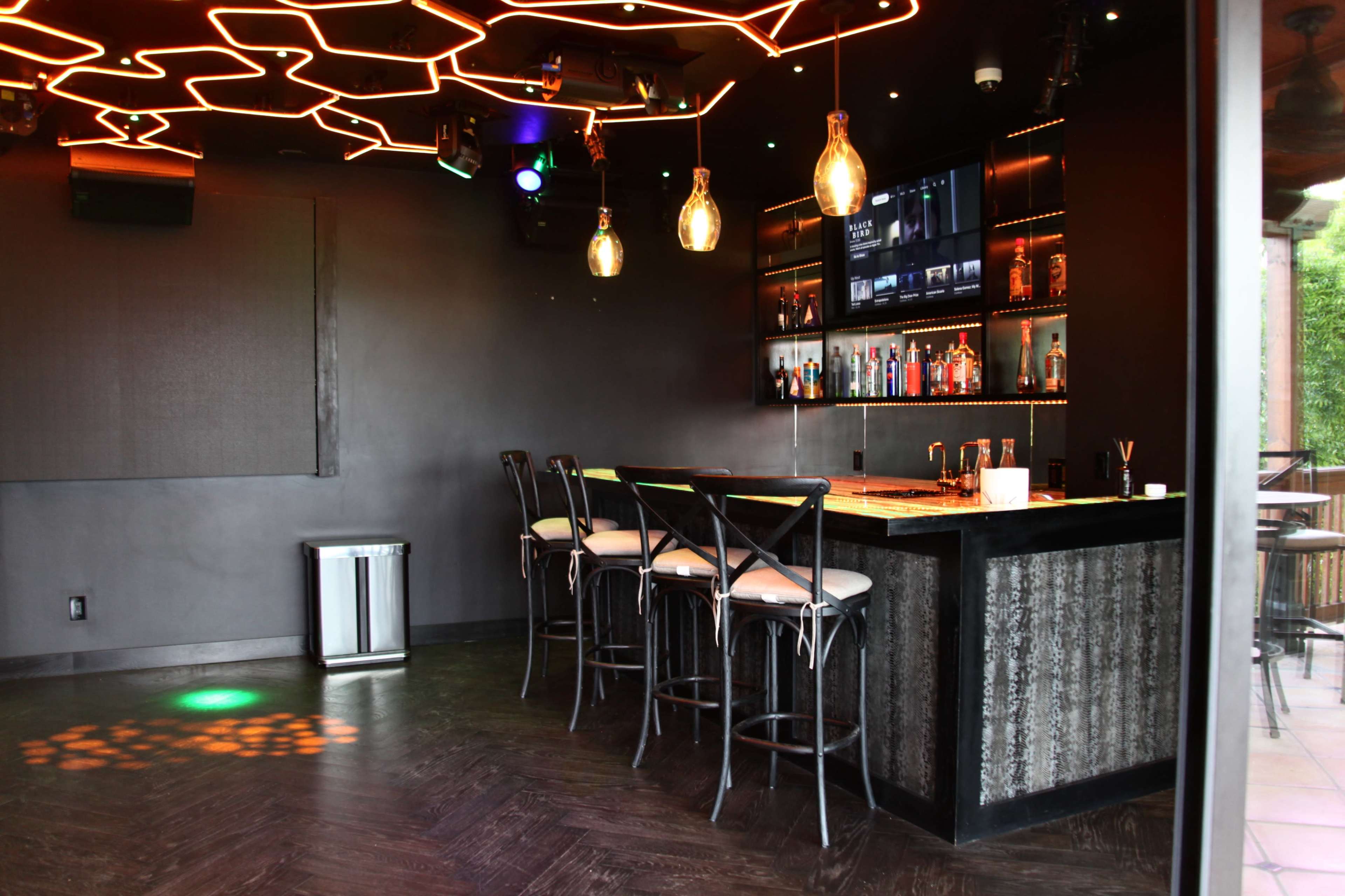 A modern bar area features a dark interior with a brightly lit ceiling design and a backlit shelf displaying various bottles.