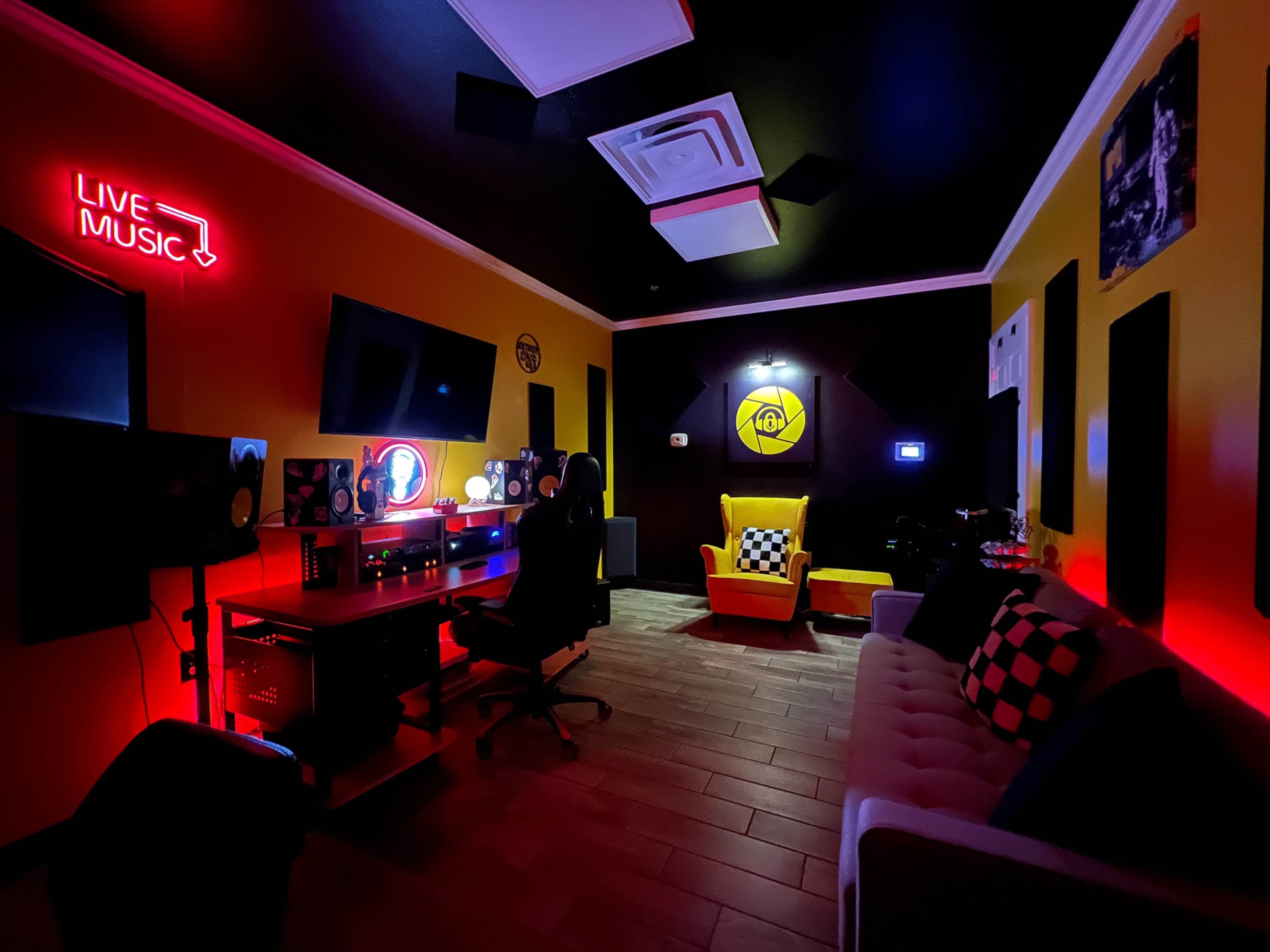 The image shows a small music recording studio furnished with a sofa, a desk, and bright colored walls featuring a "LIVE MUSIC" neon sign.