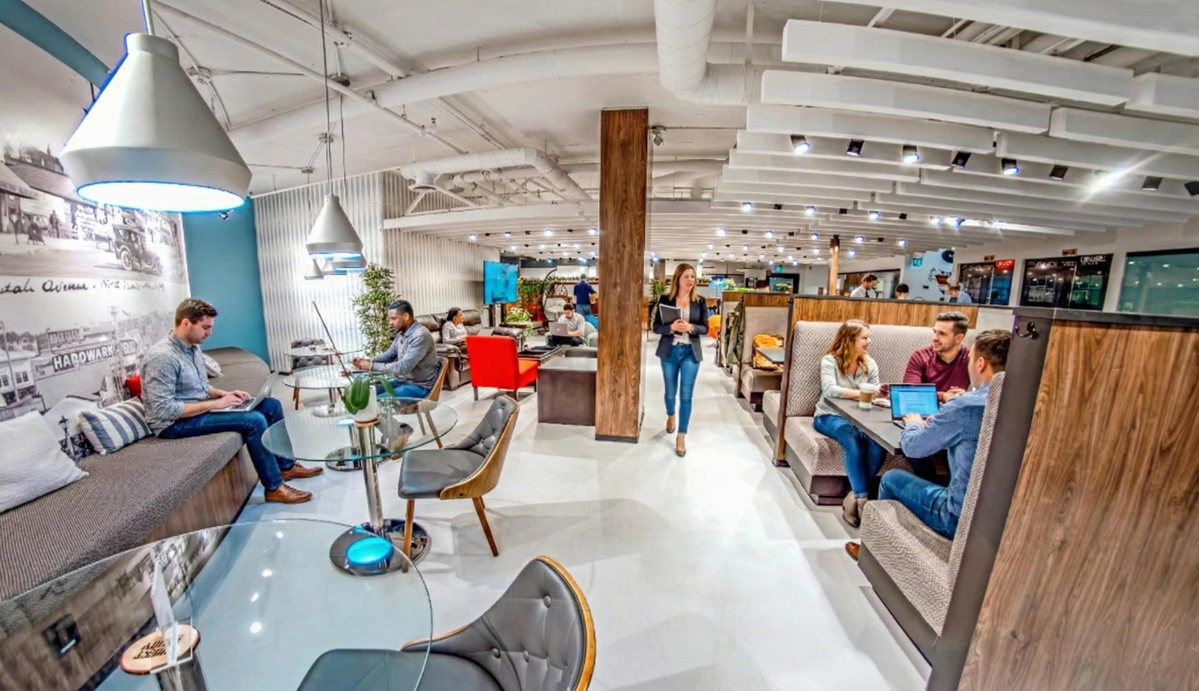 The image shows a modern co-working space with various people engaged in conversations and working on laptops amid contemporary furniture and bright lighting.