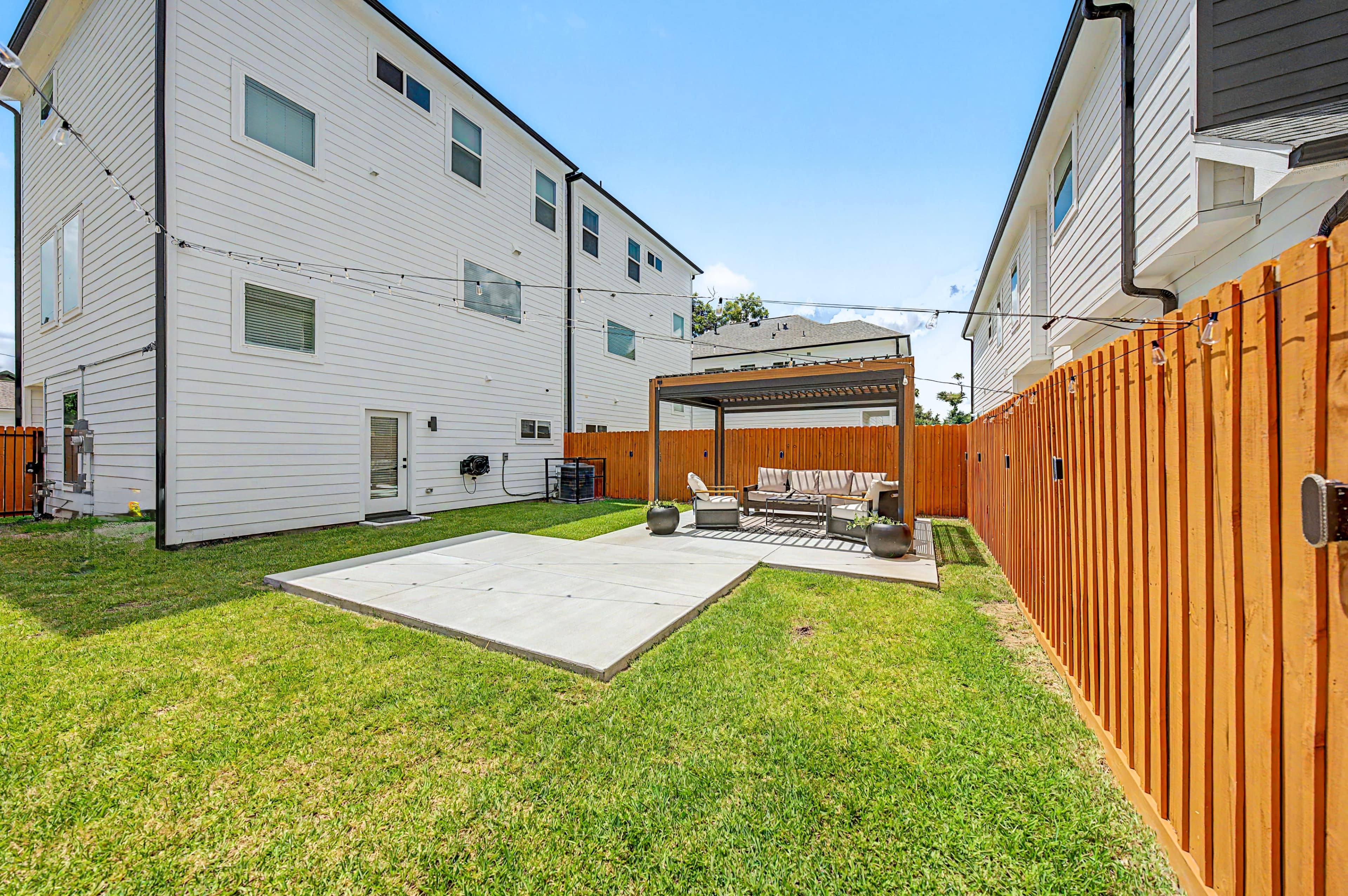 The image shows a spacious backyard featuring a concrete patio, a covered seating area, and tall wooden fencing, with two residential buildings in the background.
