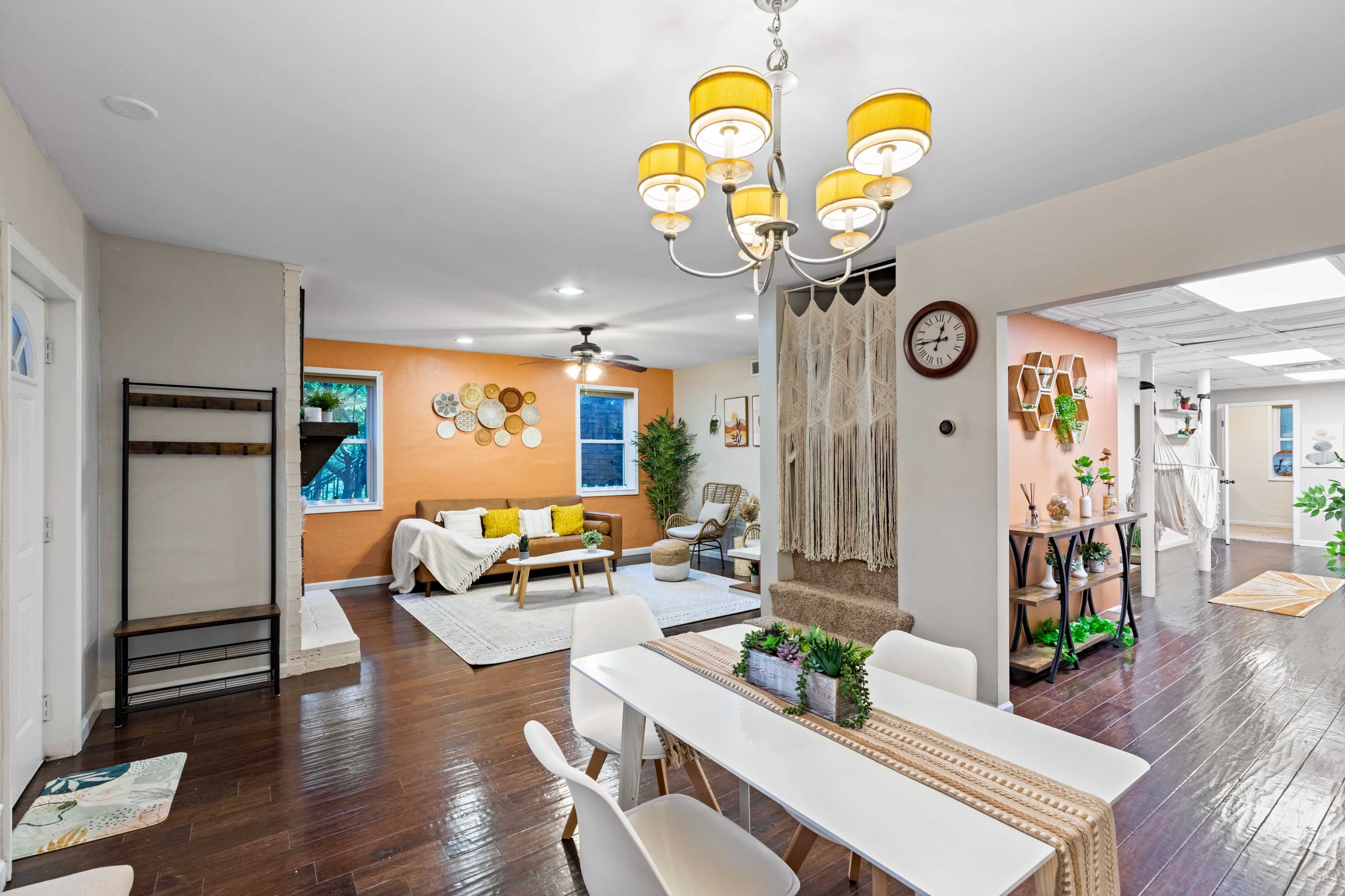 A bright living space features a dining area with a white table and chairs, an adjacent lounge with a sofa, and walls accented in orange, complemented by decorative elements.