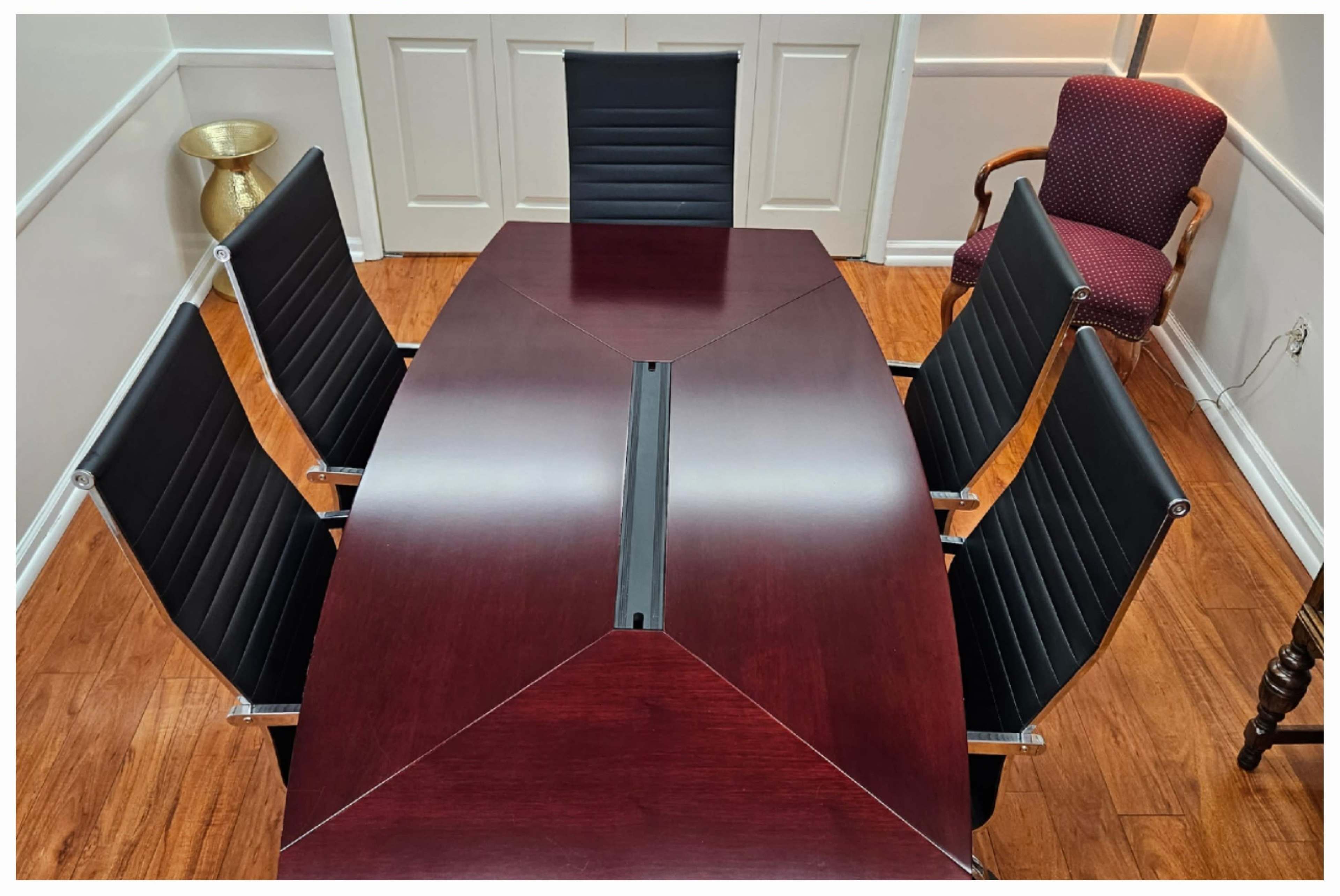 A conference room table with a polished wooden surface is surrounded by five black leather chairs, set in a well-lit space with a wall-mounted light and an accent chair in the corner.
