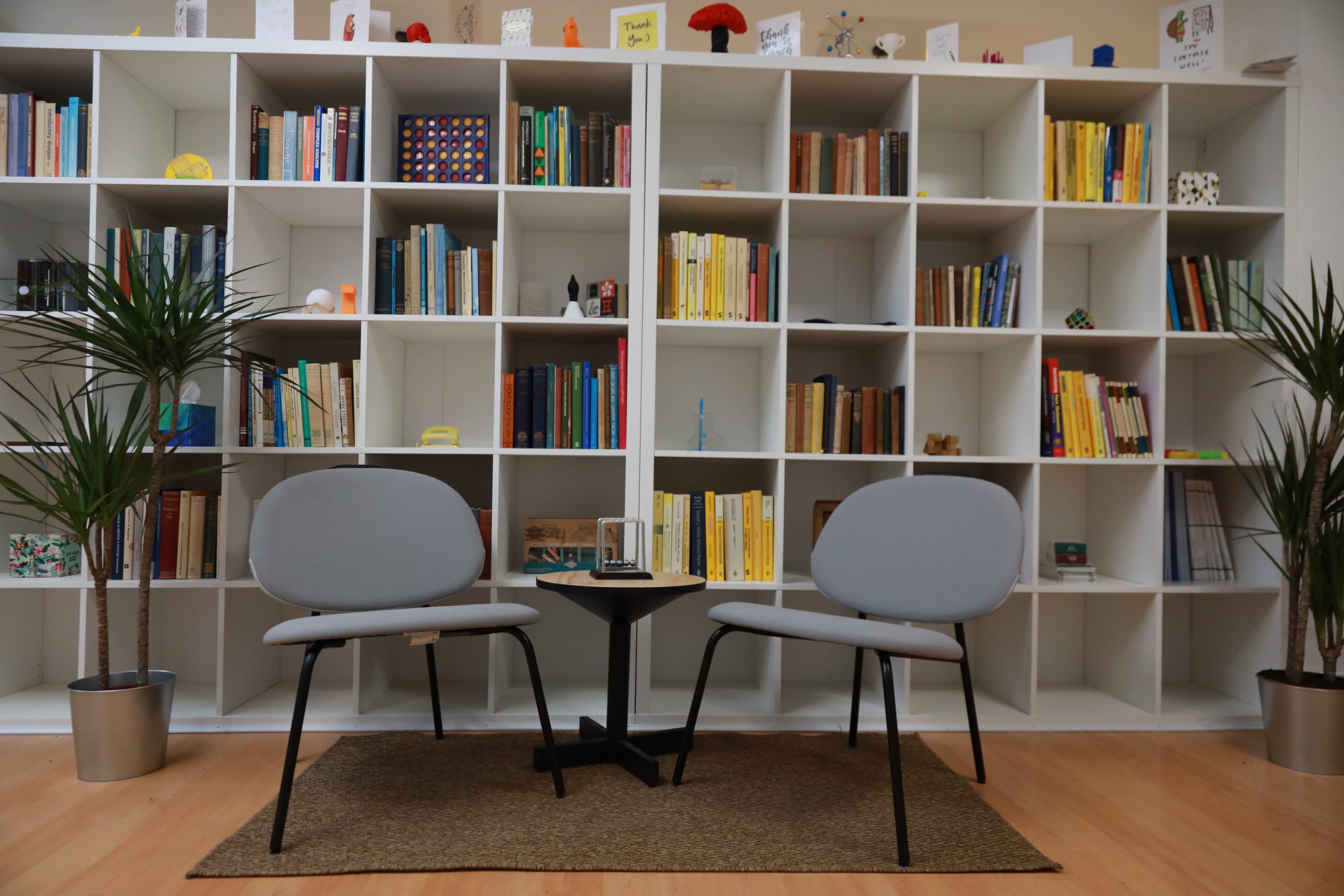 The image shows a cozy reading area with two gray chairs facing a small black table, set against a backdrop of neatly organized bookshelves filled with books and decorative items.
