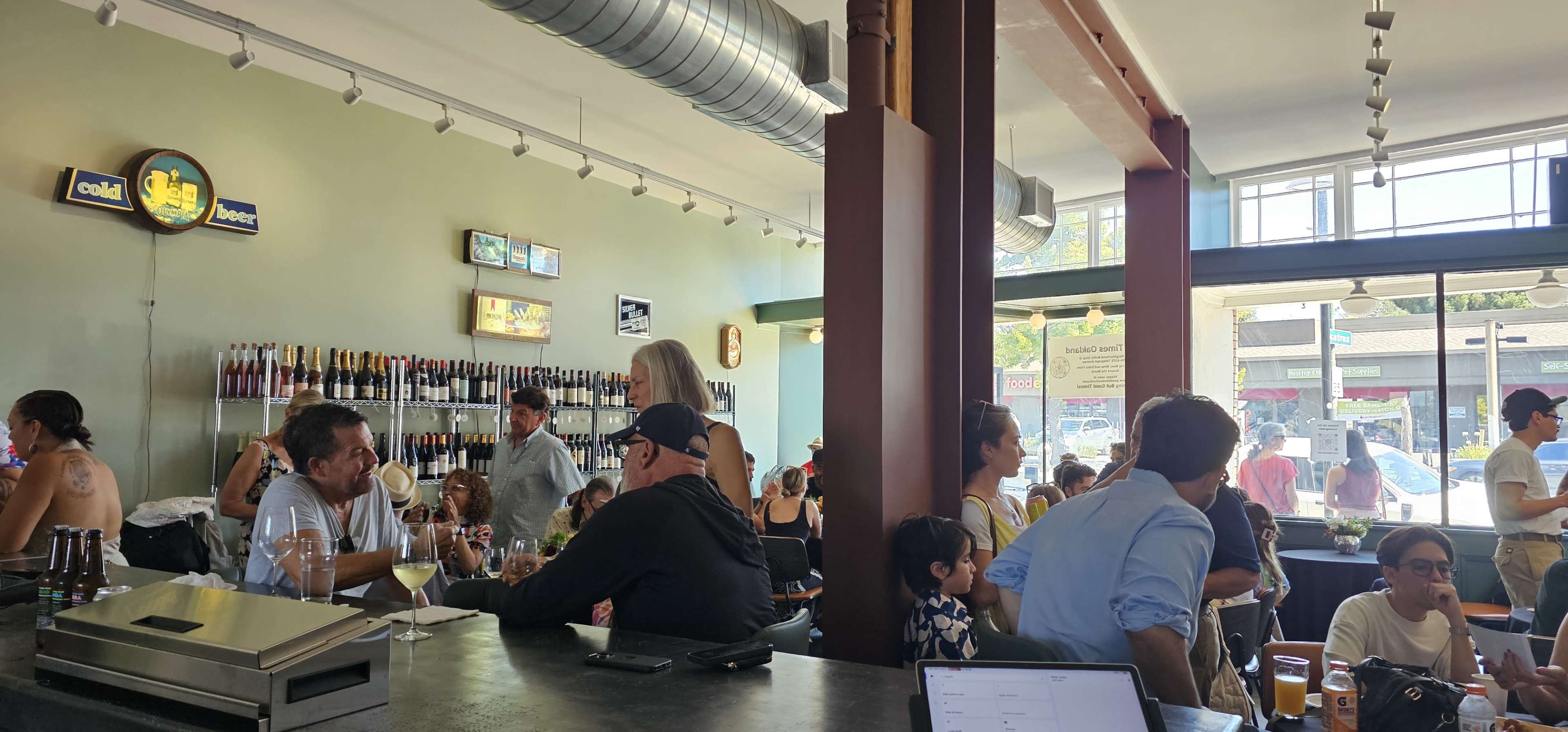 A busy bar scene shows patrons seated and standing around the counter, socializing while a wall behind them displays various bottles and beers.