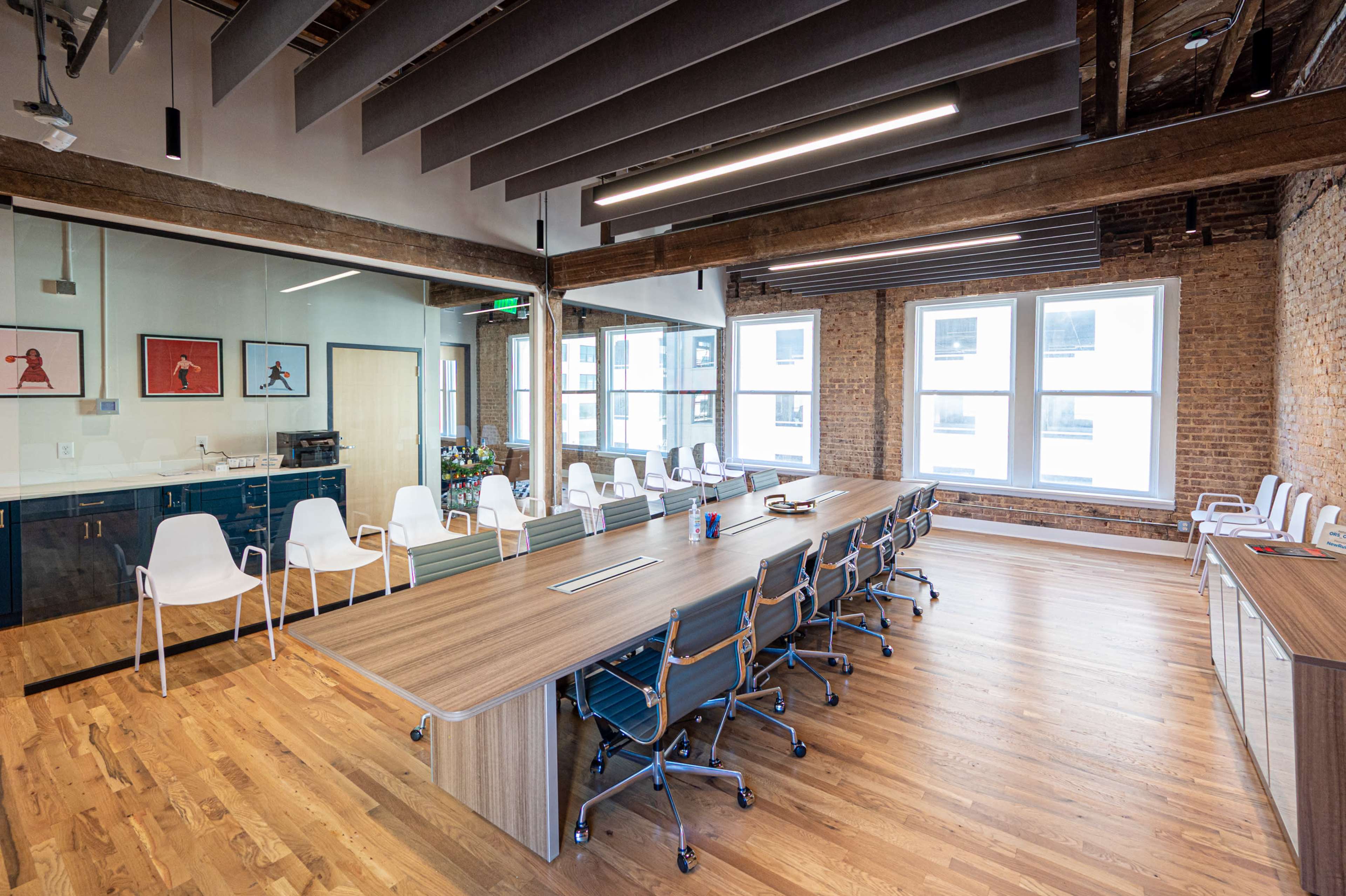 A modern conference room featuring a large wooden table surrounded by ergonomic chairs, with large windows and exposed brick walls.