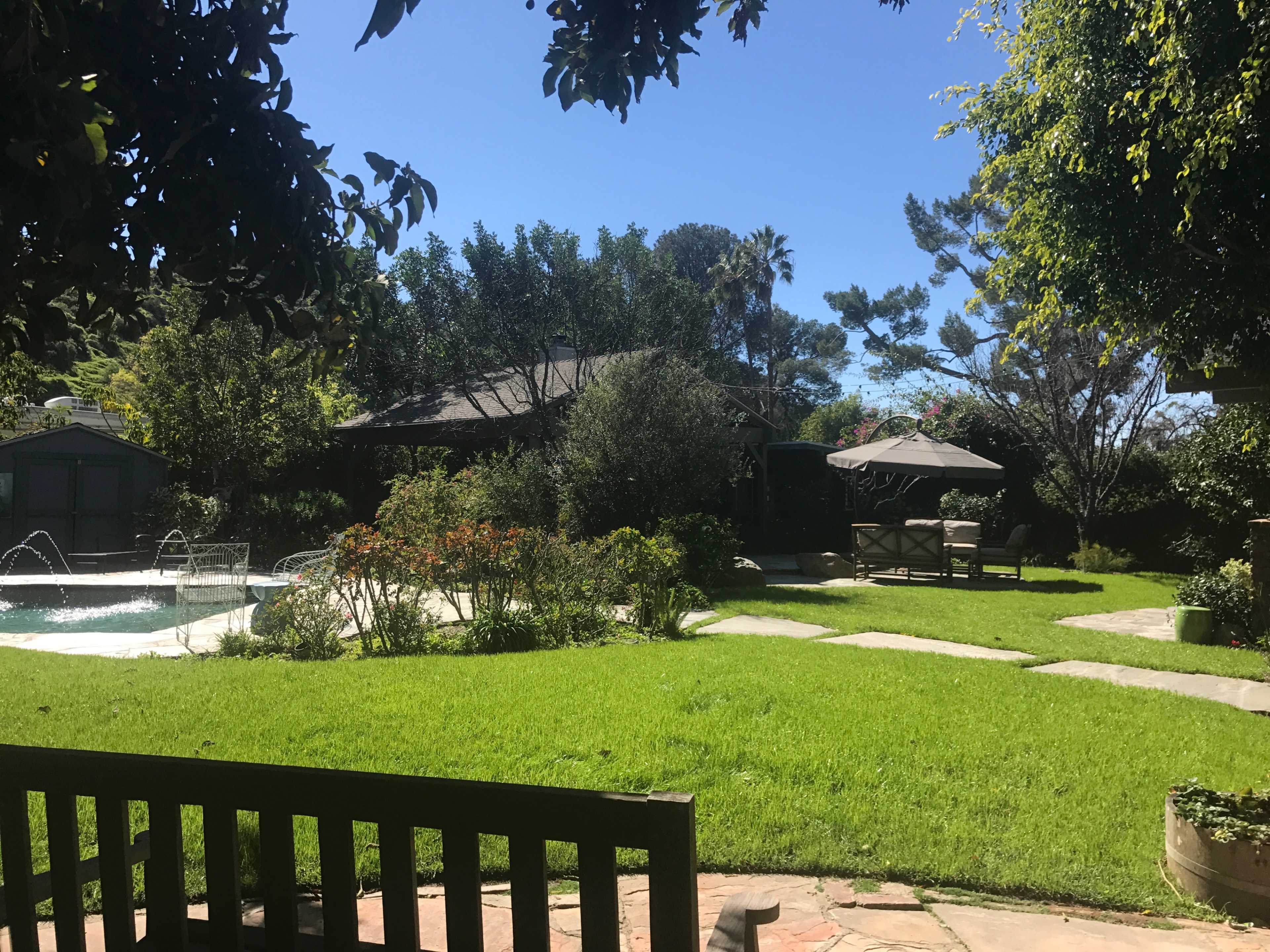 The image shows a landscaped garden with a swimming pool, patio seating, and various trees and plants under a clear blue sky.