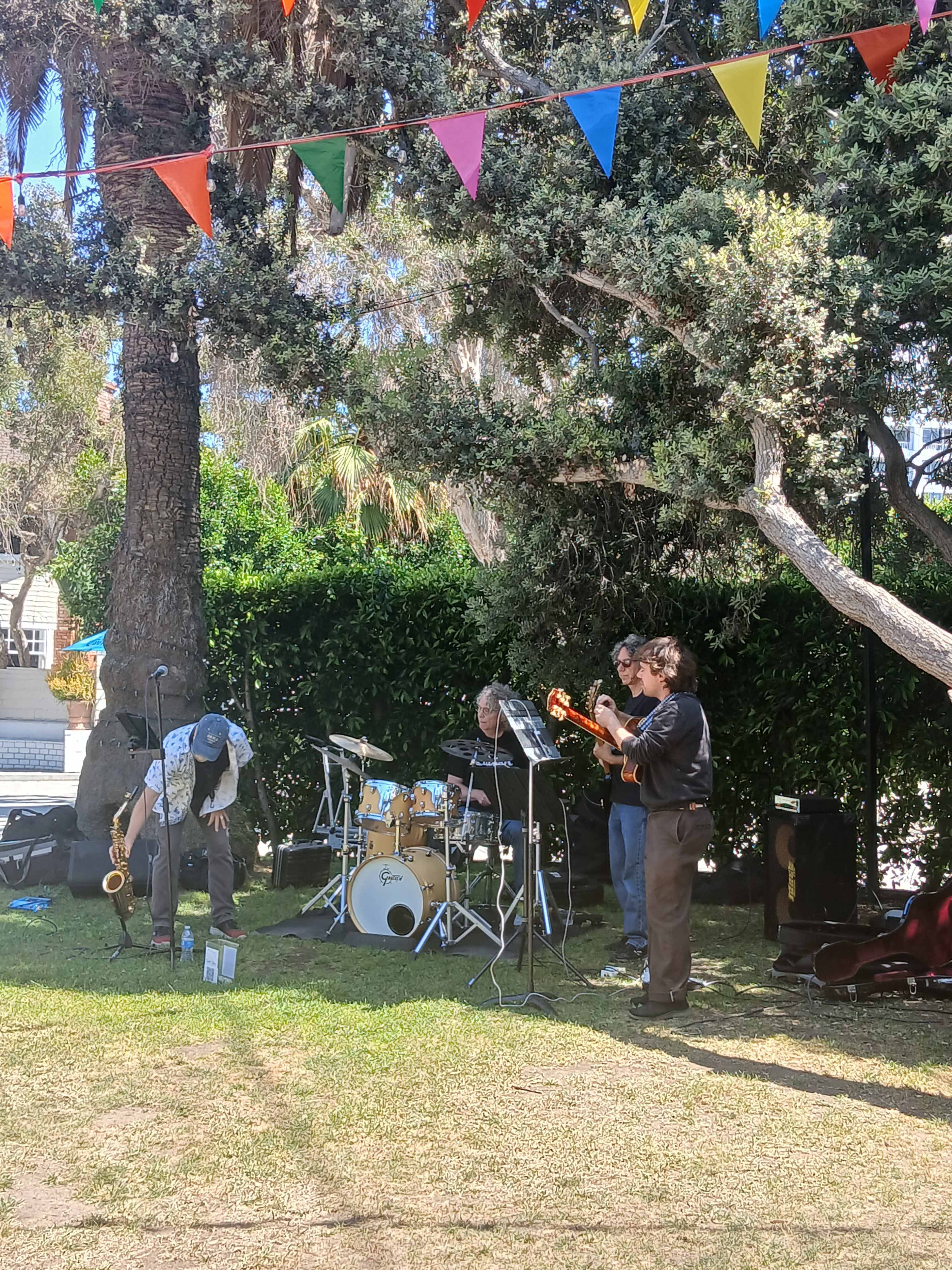 A small band performs on a grassy area, with colorful banners hanging overhead and a tree in the background.