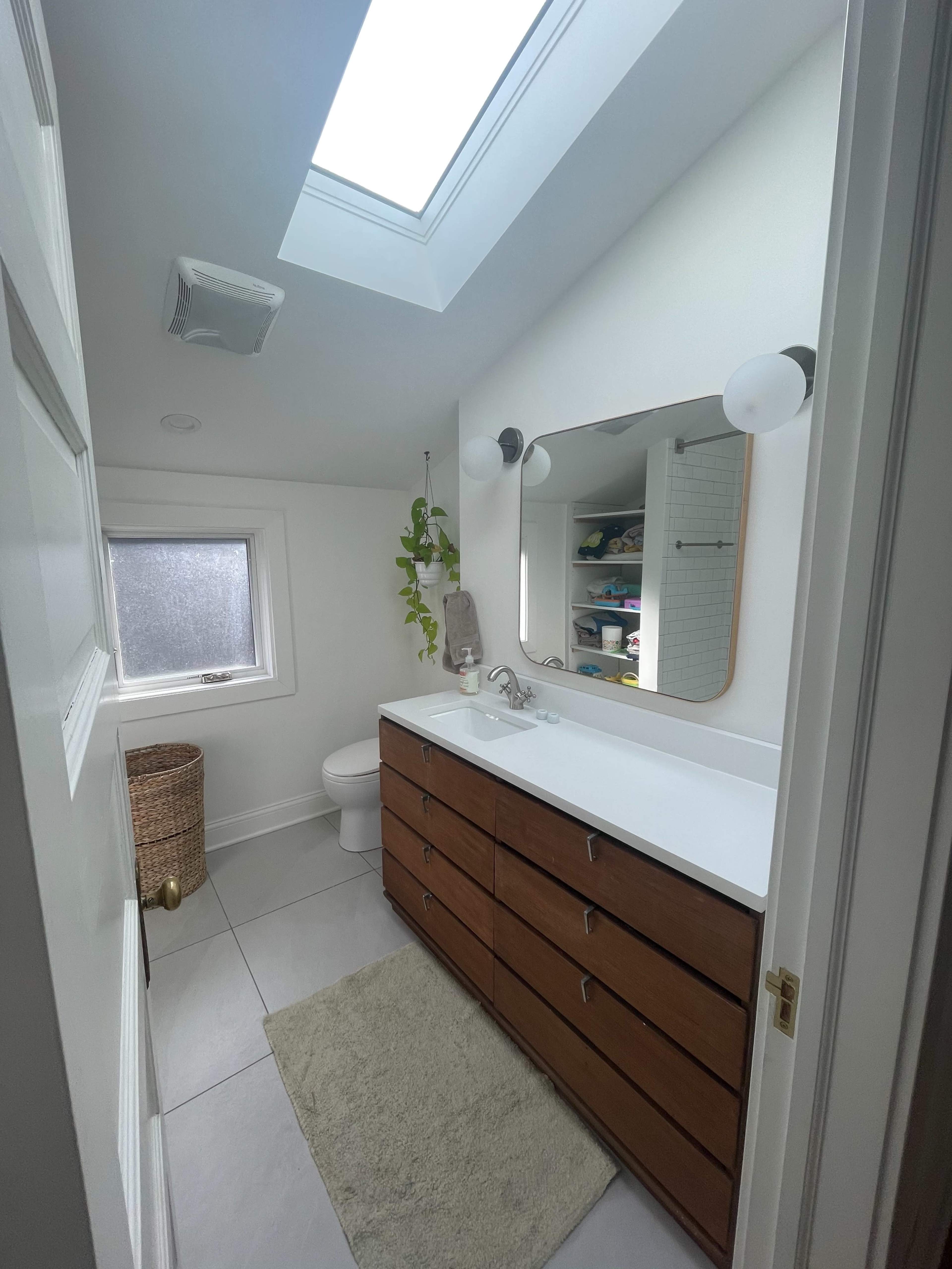 A modern bathroom featuring a wooden vanity with multiple drawers, a skylight, and a window, along with a toilet and a small basket.