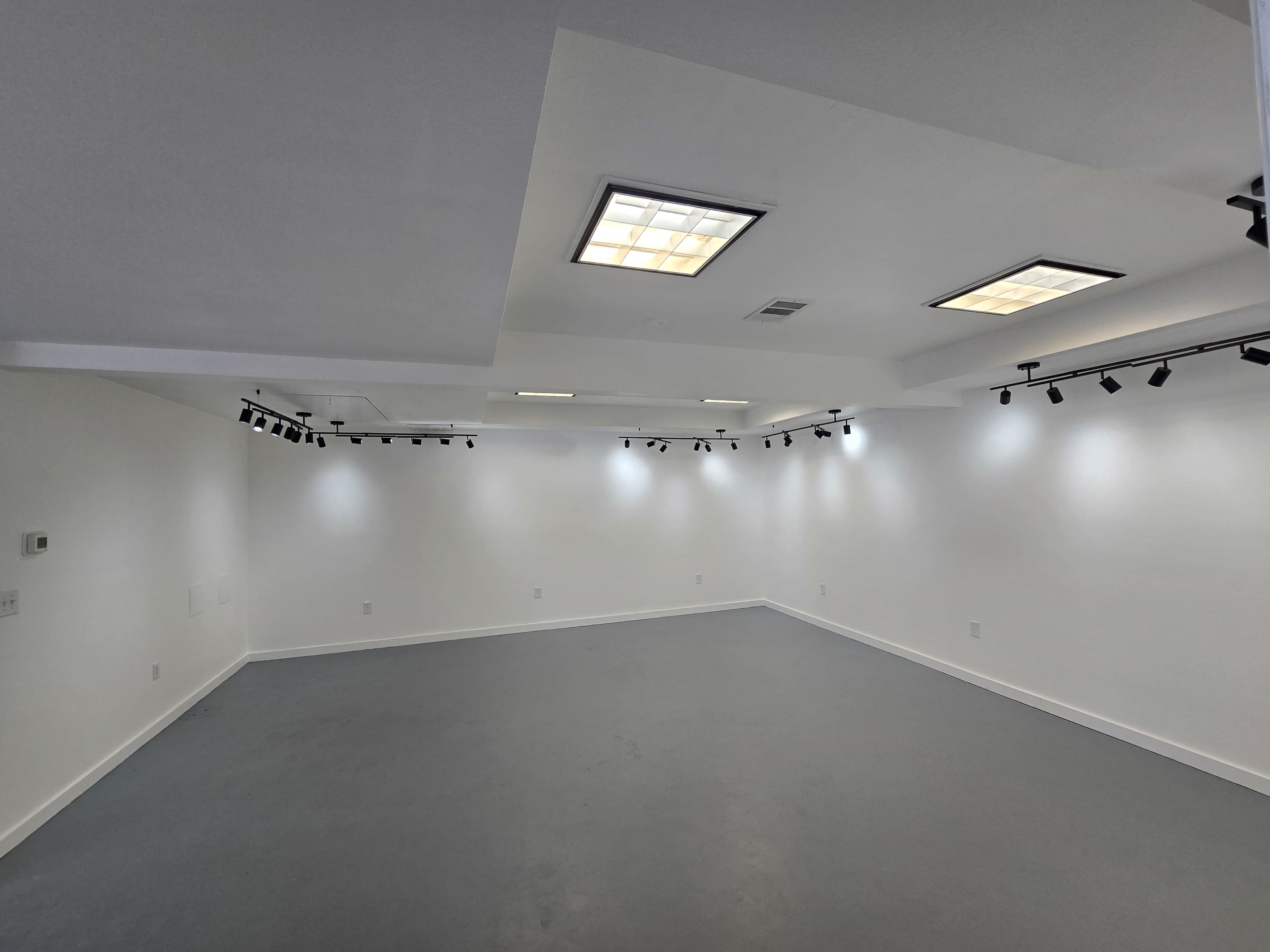 The image shows a large, empty room with white walls and a grey floor, featuring recessed lighting and track lights.