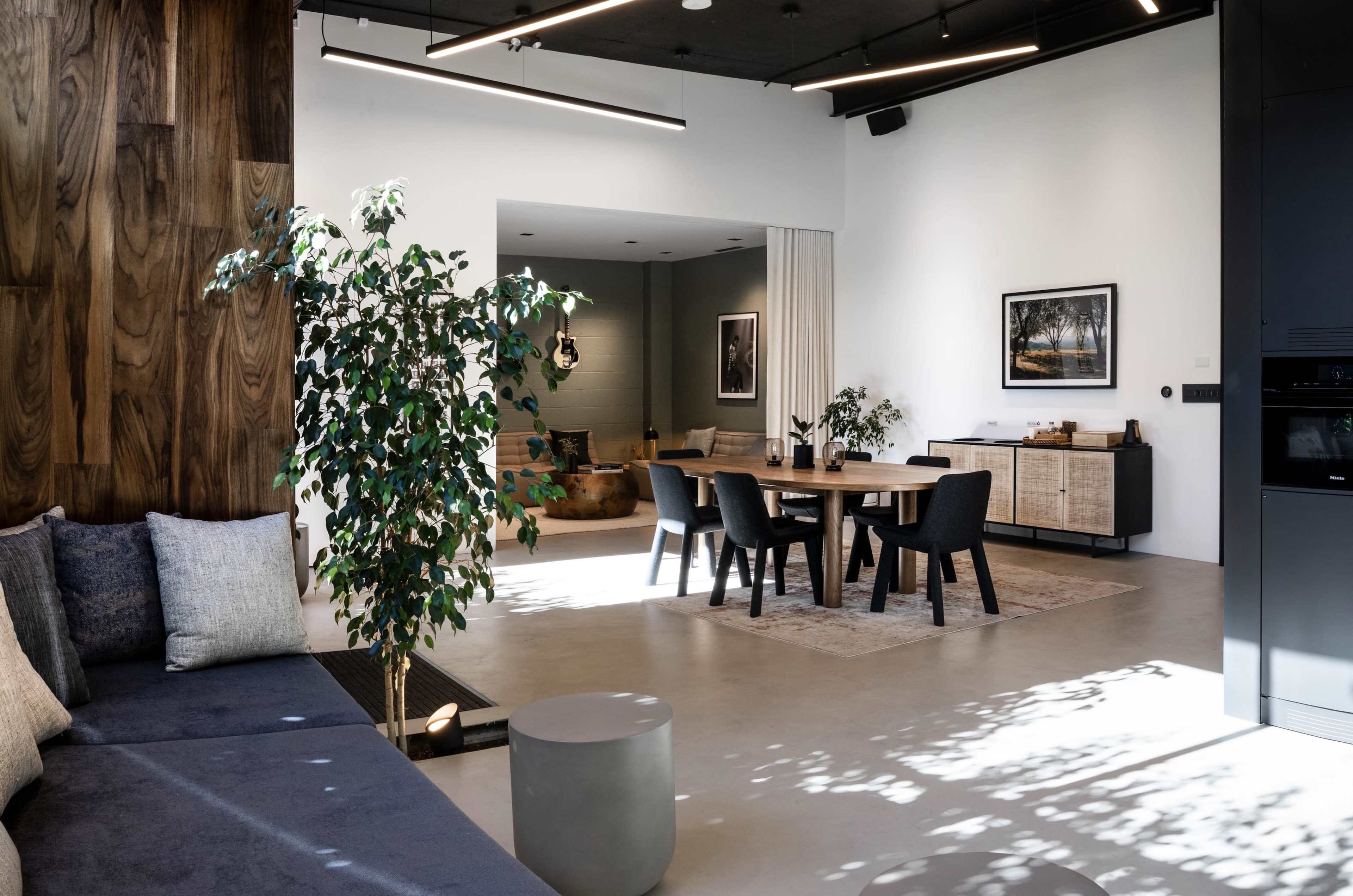 The image shows a modern, open-plan living space featuring a flexible seating area, a dining table with chairs, and decorative plants.