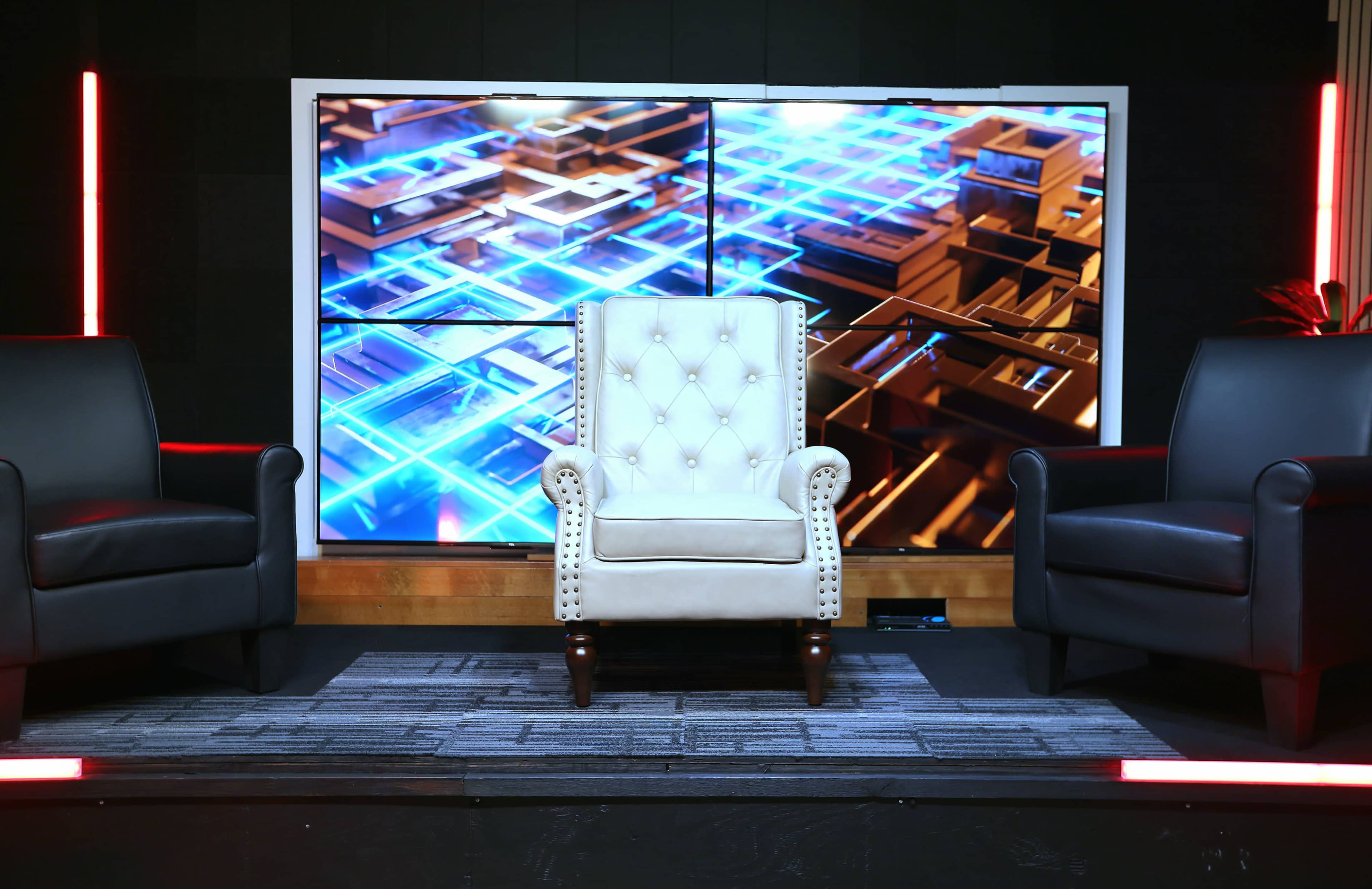 A central tufted white armchair is flanked by two black armchairs in front of a large screen displaying a futuristic digital design.