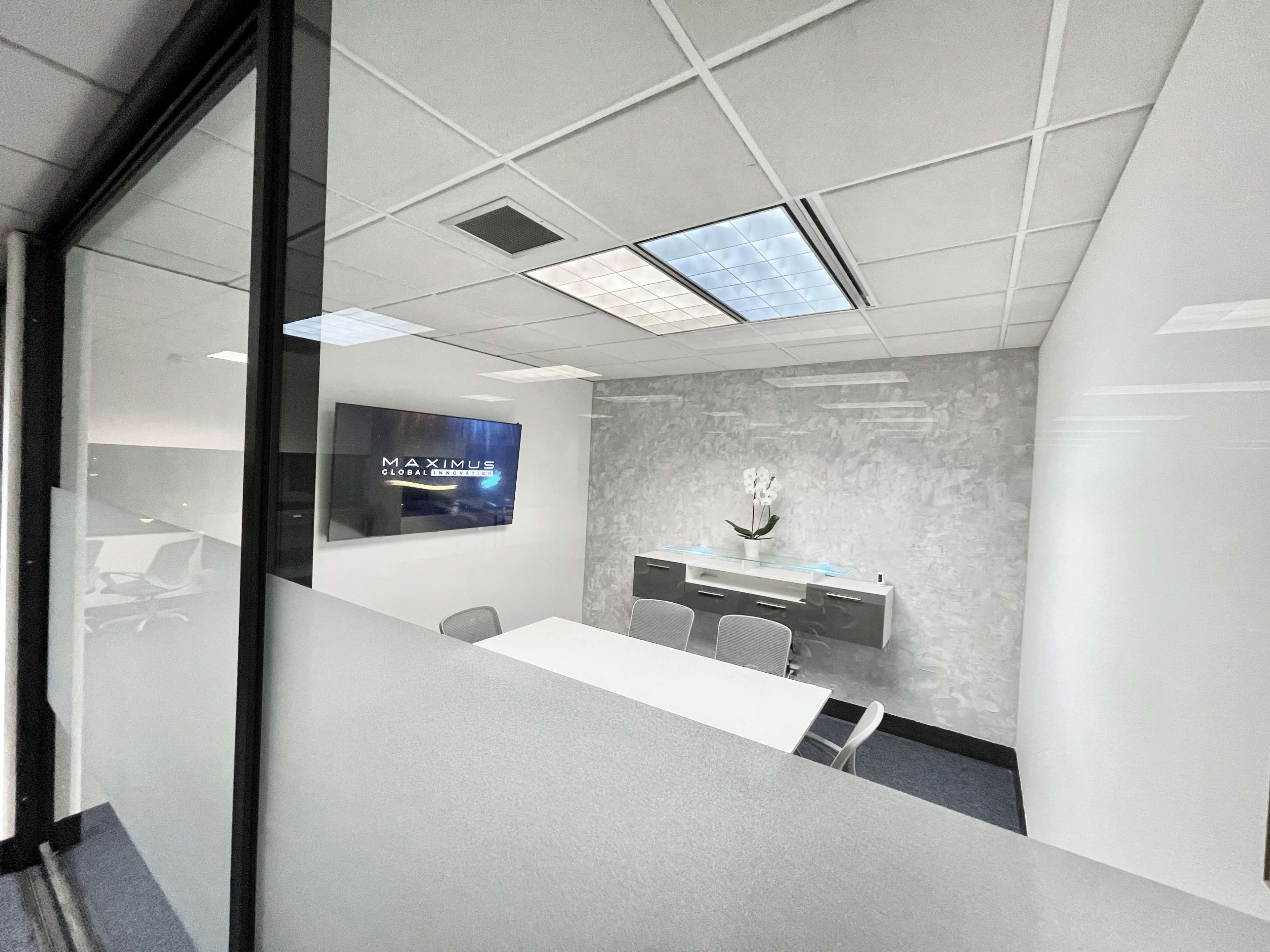 The image shows a modern office meeting room with a glass wall, featuring a white table, gray chairs, and a wall-mounted screen displaying the logo "MAXIMUS."