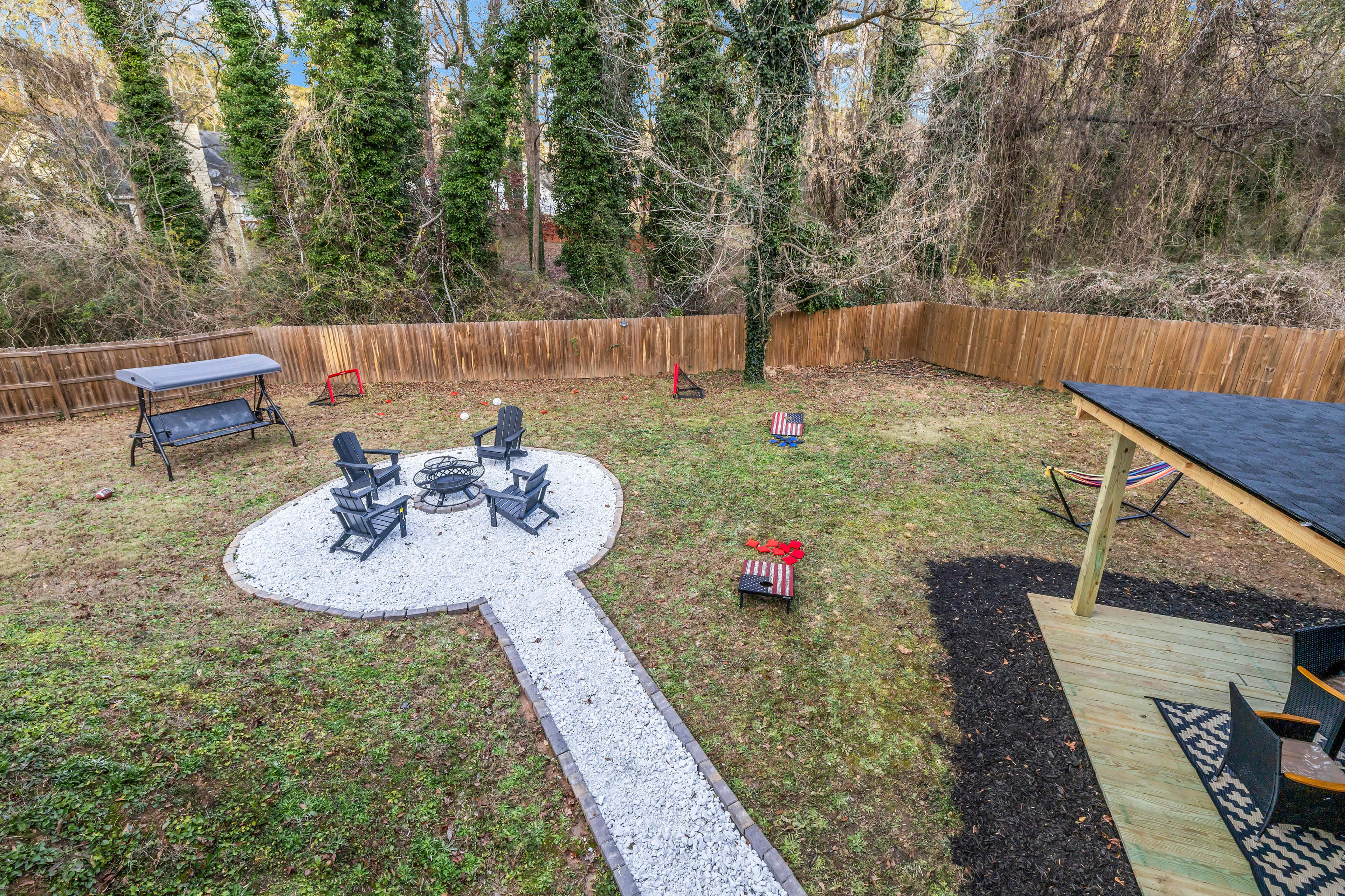 The image shows a backyard with a stone pathway leading to a fire pit area surrounded by four chairs, framed by a wooden fence and patches of grass.