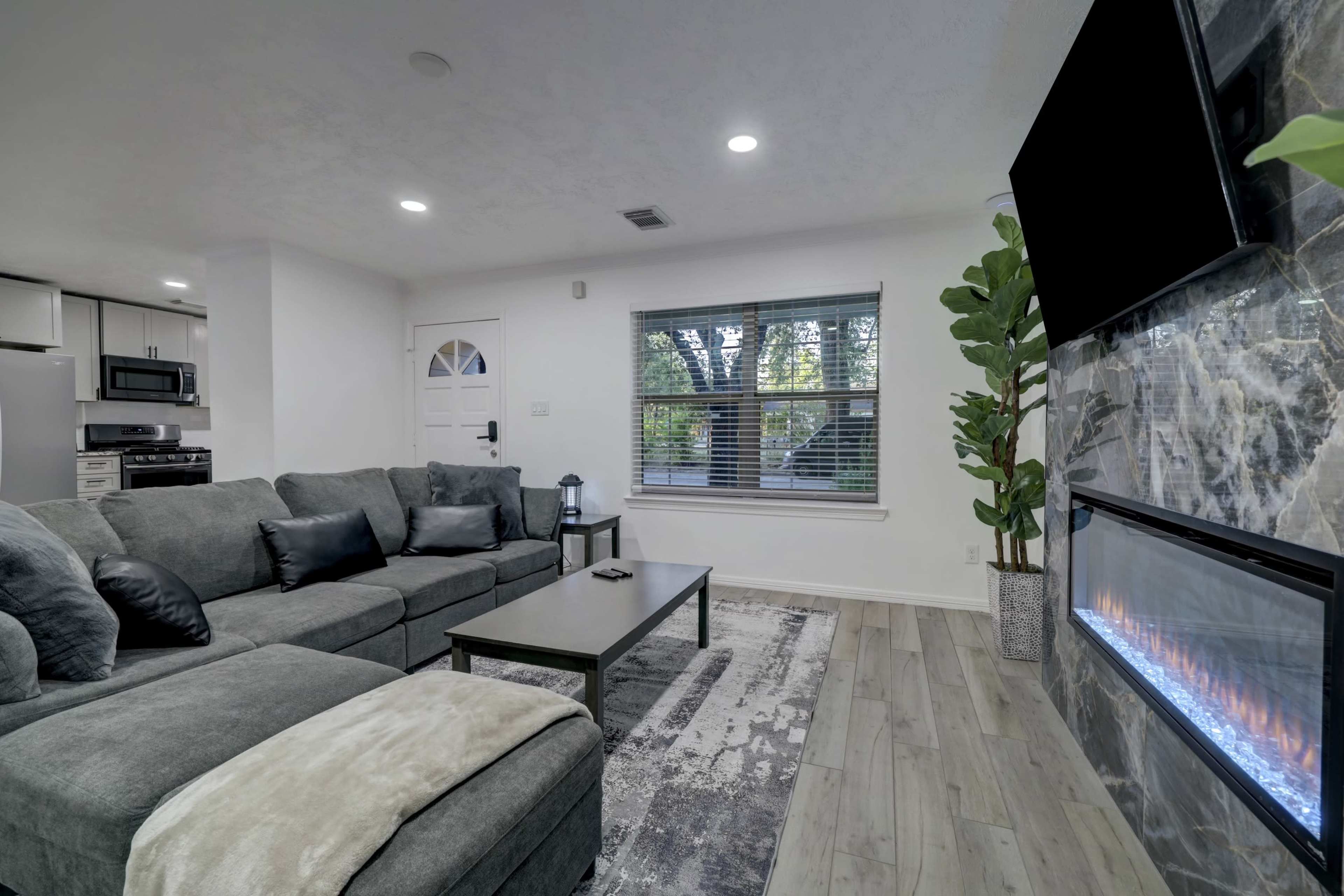 A modern living room featuring a gray sectional sofa, a coffee table, an electric fireplace, and a large window with view of greenery outside.