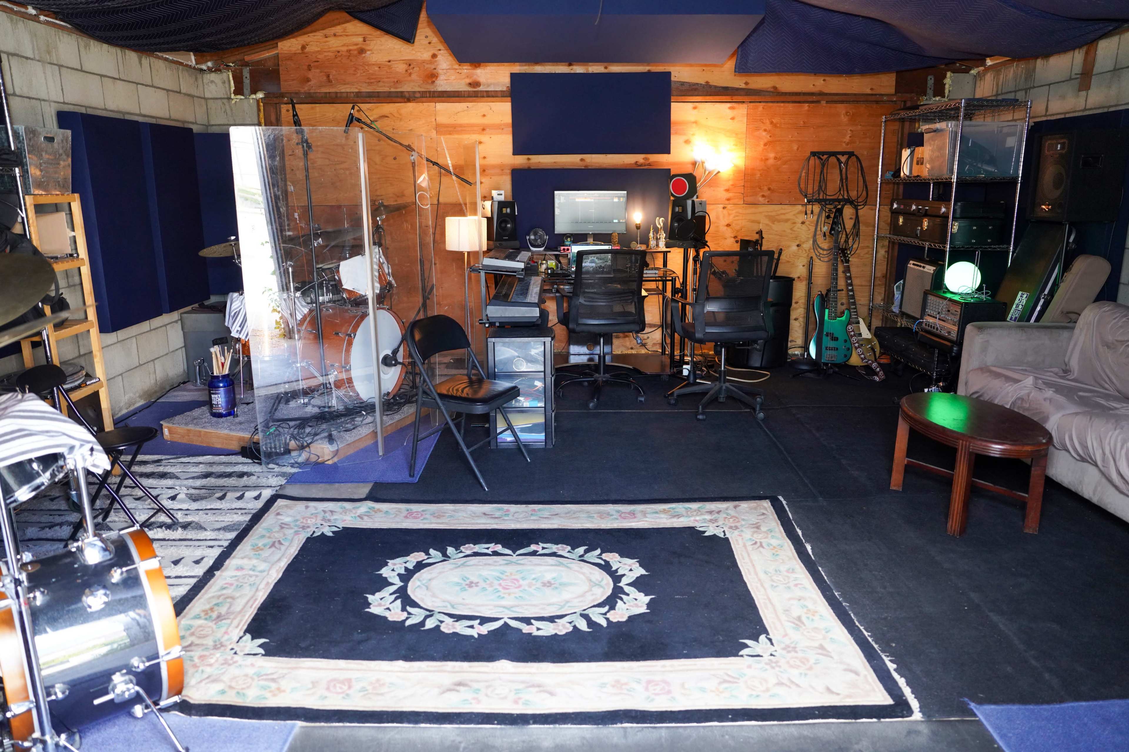 The image shows a music studio with a drum set, two office chairs, a computer setup, and various musical instruments arranged in a wood-paneled space.