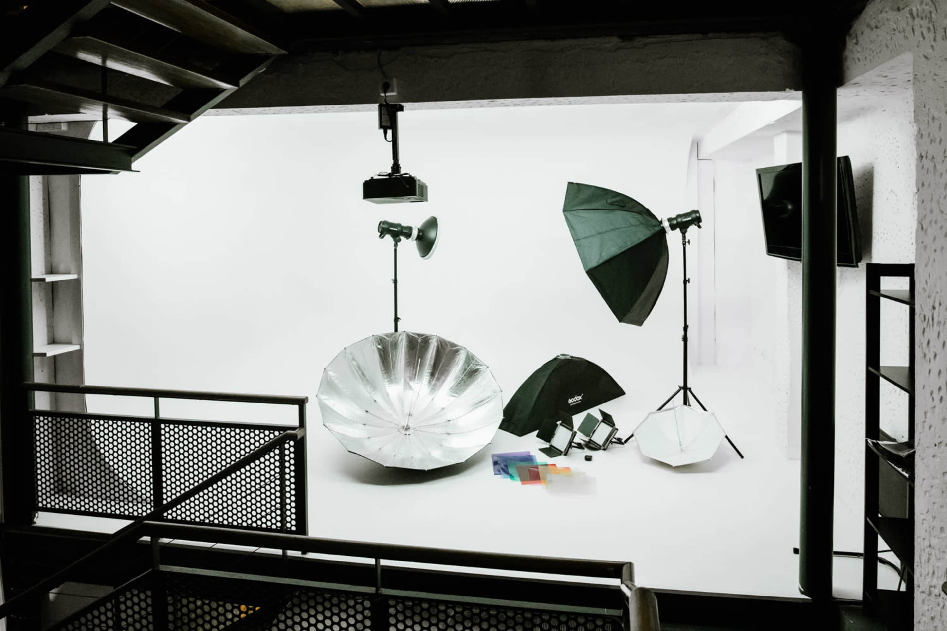 A photography studio setup is shown with various lighting equipment, including umbrellas and softboxes, arranged against a white backdrop.