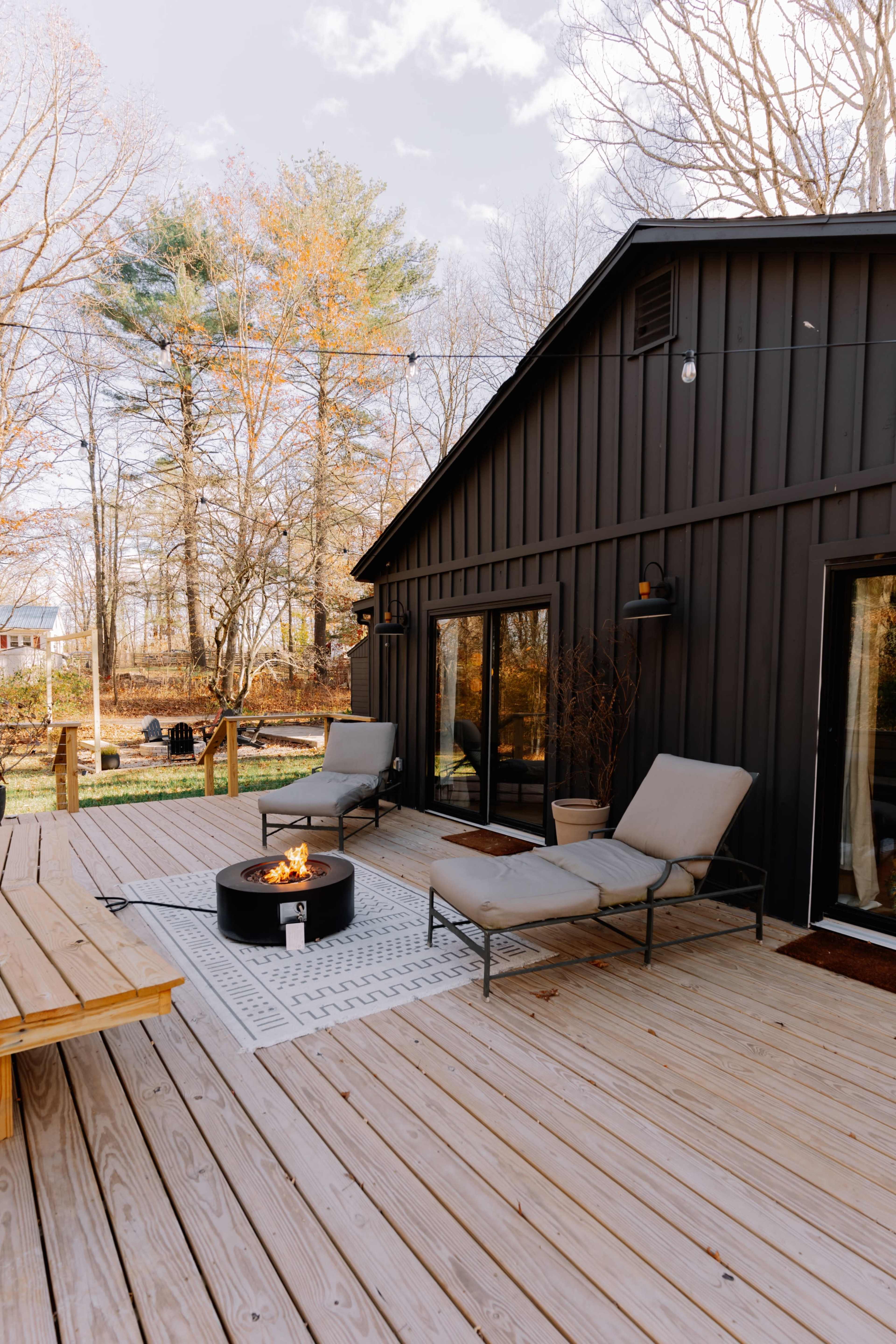 A wooden deck features two lounge chairs beside a circular fire pit, in a wooded outdoor setting.