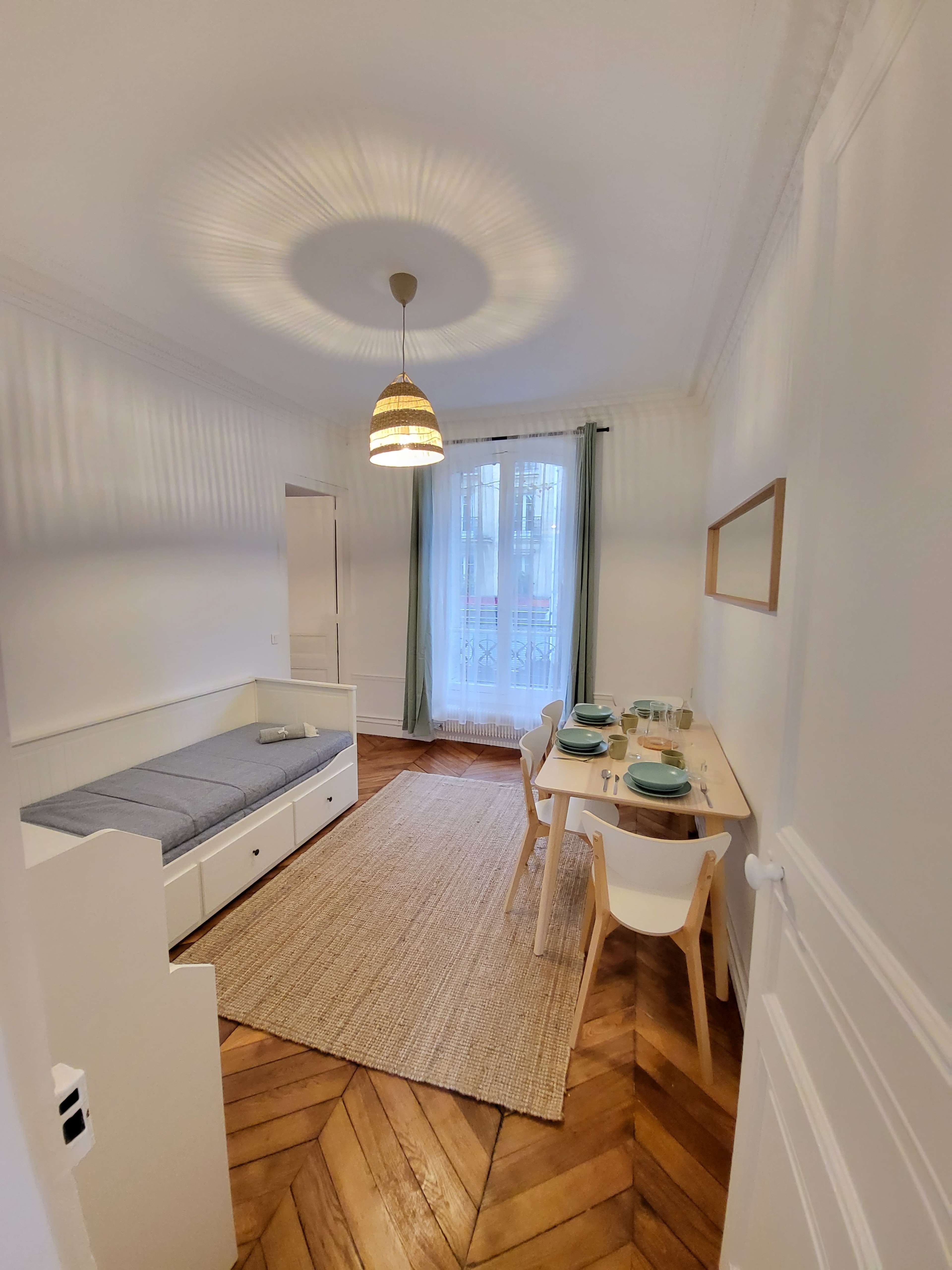 The image shows a brightly lit, minimalistic room featuring a bed, a dining table set for two, and a large window with light curtains.