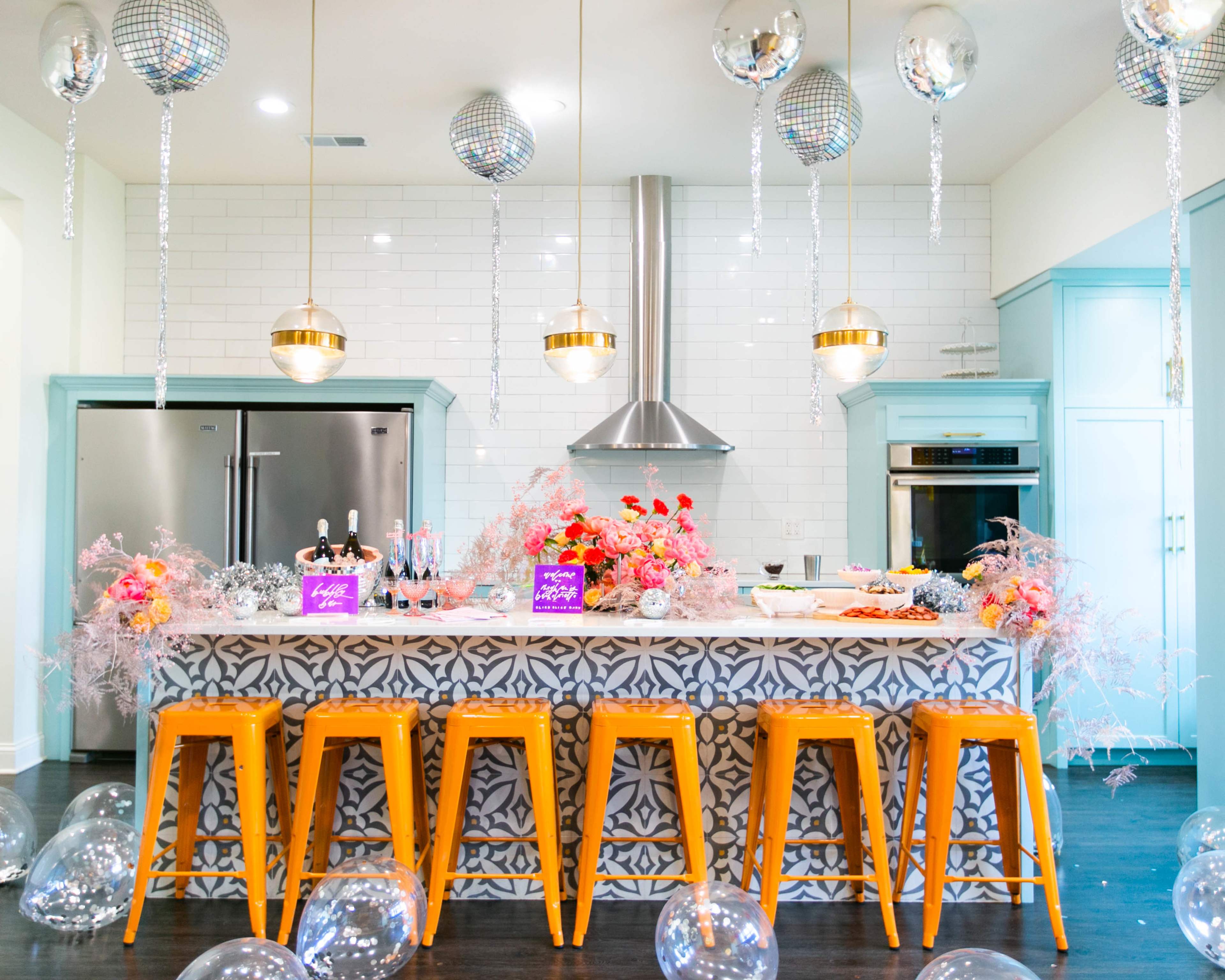 A modern kitchen features a vibrant bar with orange stools, a marble countertop adorned with floral decorations, and silver disco balls hanging from the ceiling.