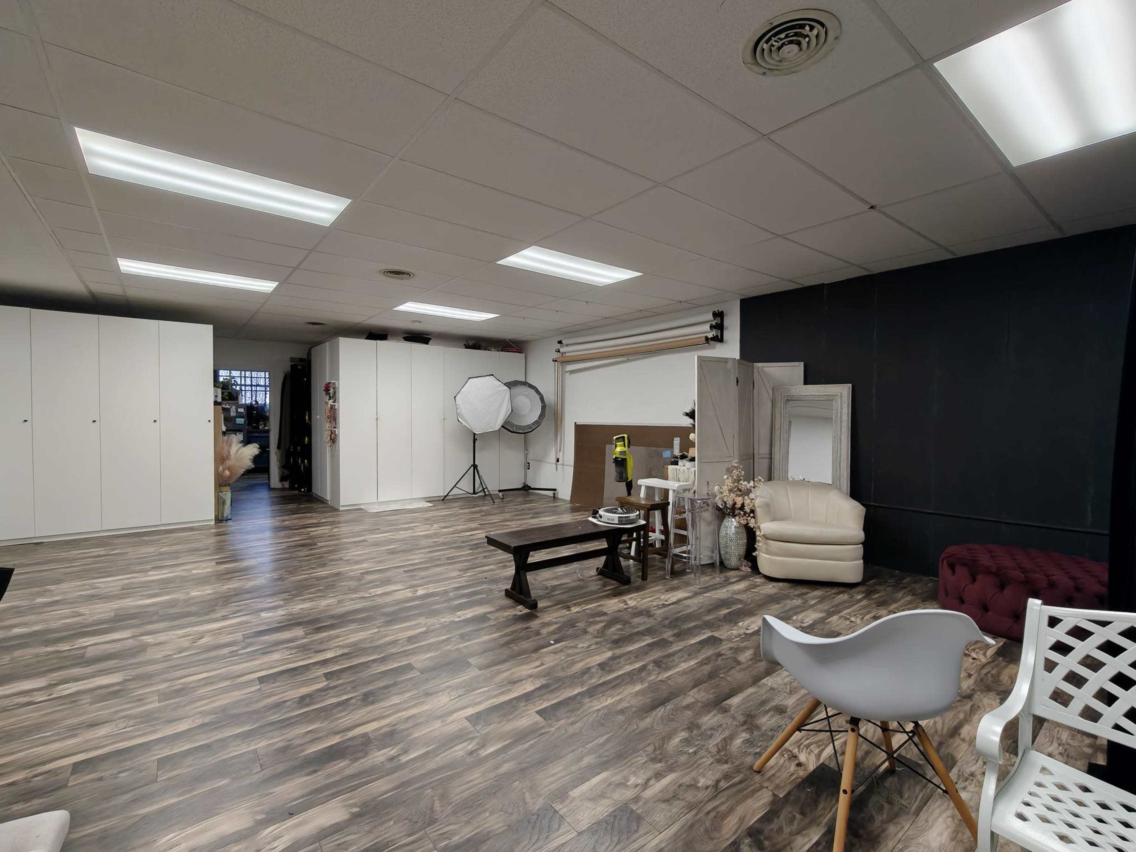 The image shows a spacious room with wood-patterned flooring, featuring various furniture pieces, lighting equipment, and storage cabinets against the walls.